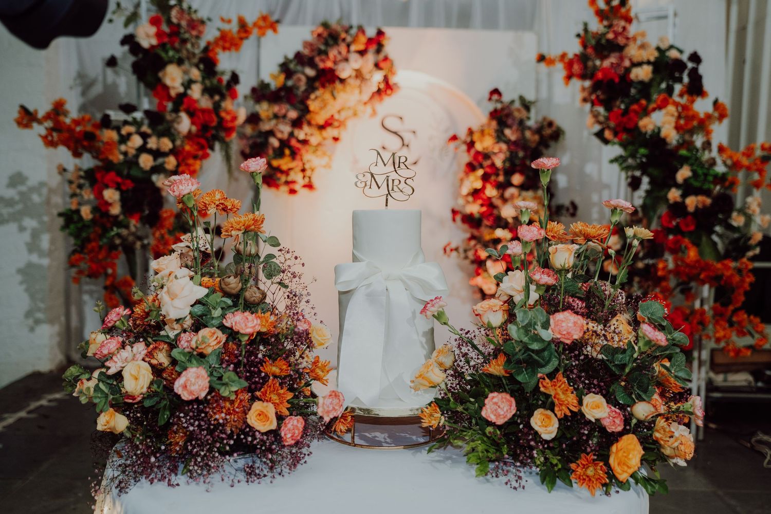 Wedding stage with a white cake and name sign surrounded by orange, pink and red floral arrangements.