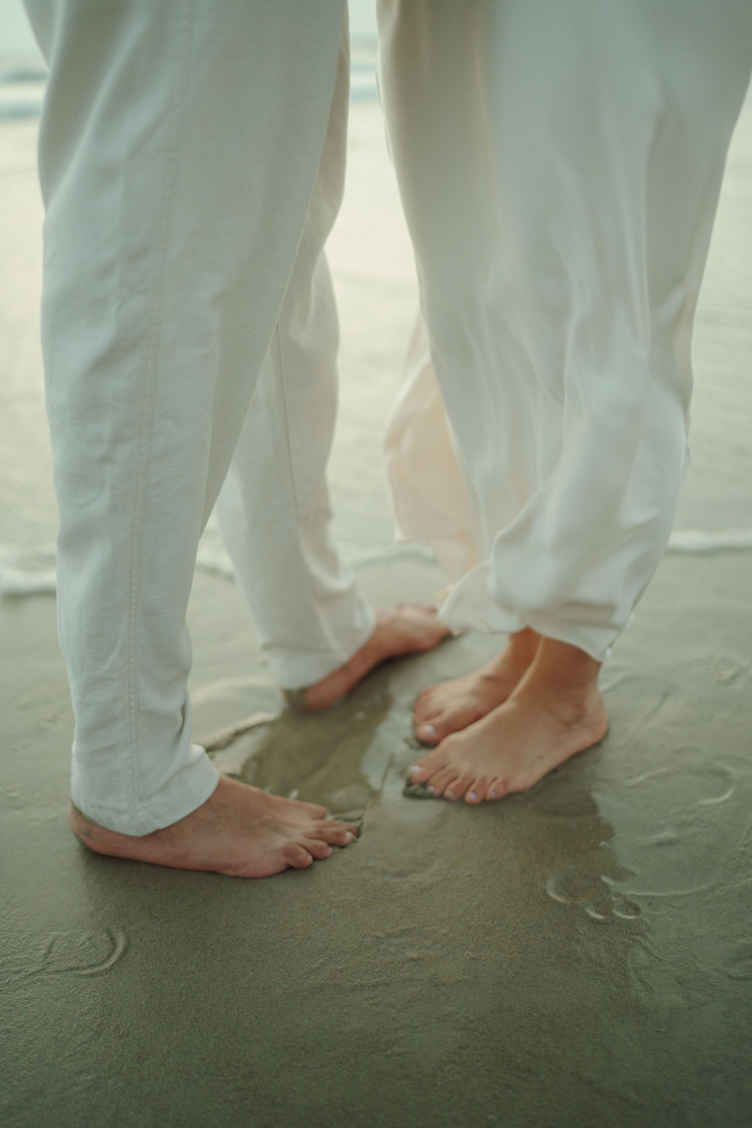 Two people in white linen pants stand barefoot on wet sand at the beach shoreline.