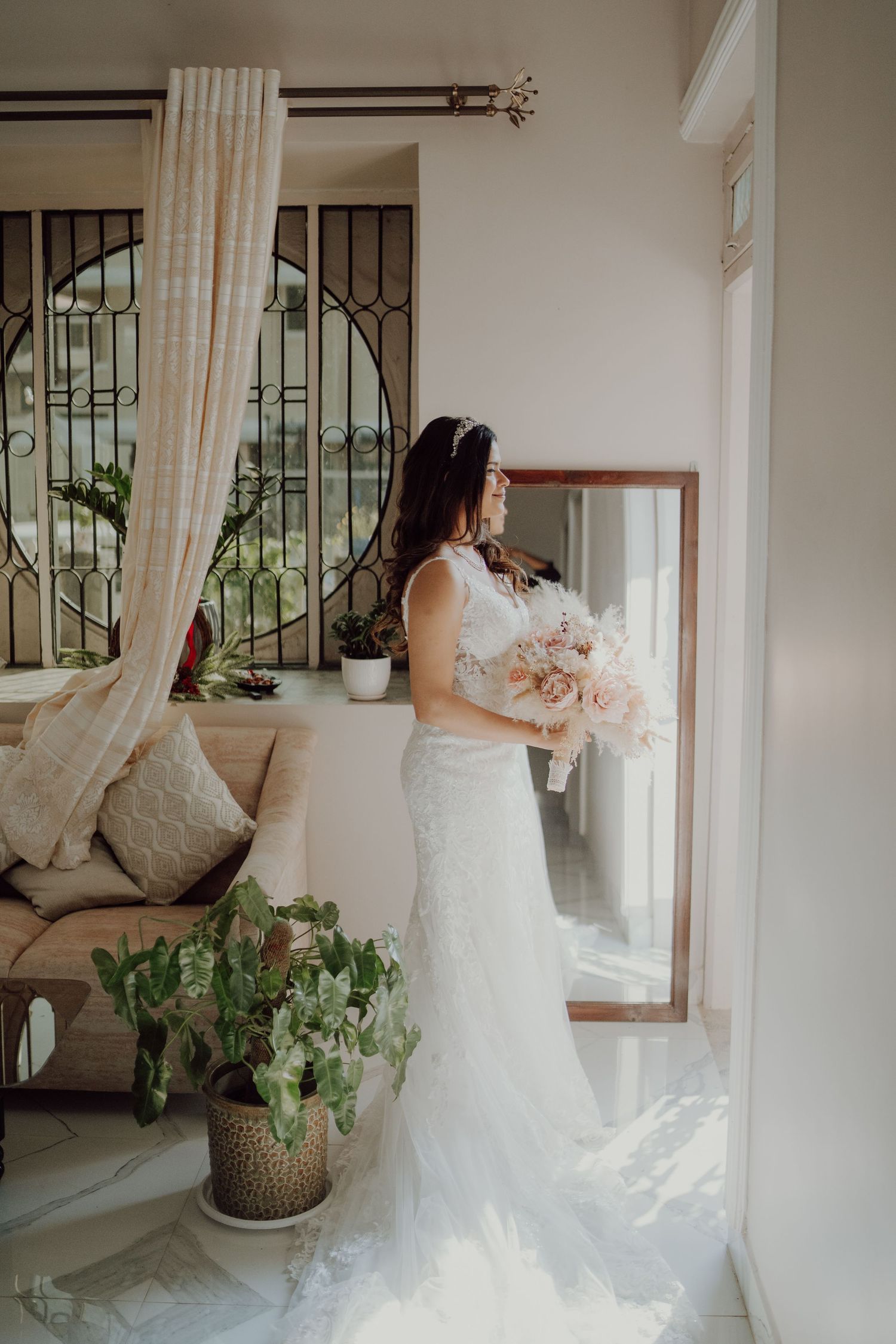 Elegant bridal portrait showing full-length lace gown and bouquet beside window with decorative iron details.