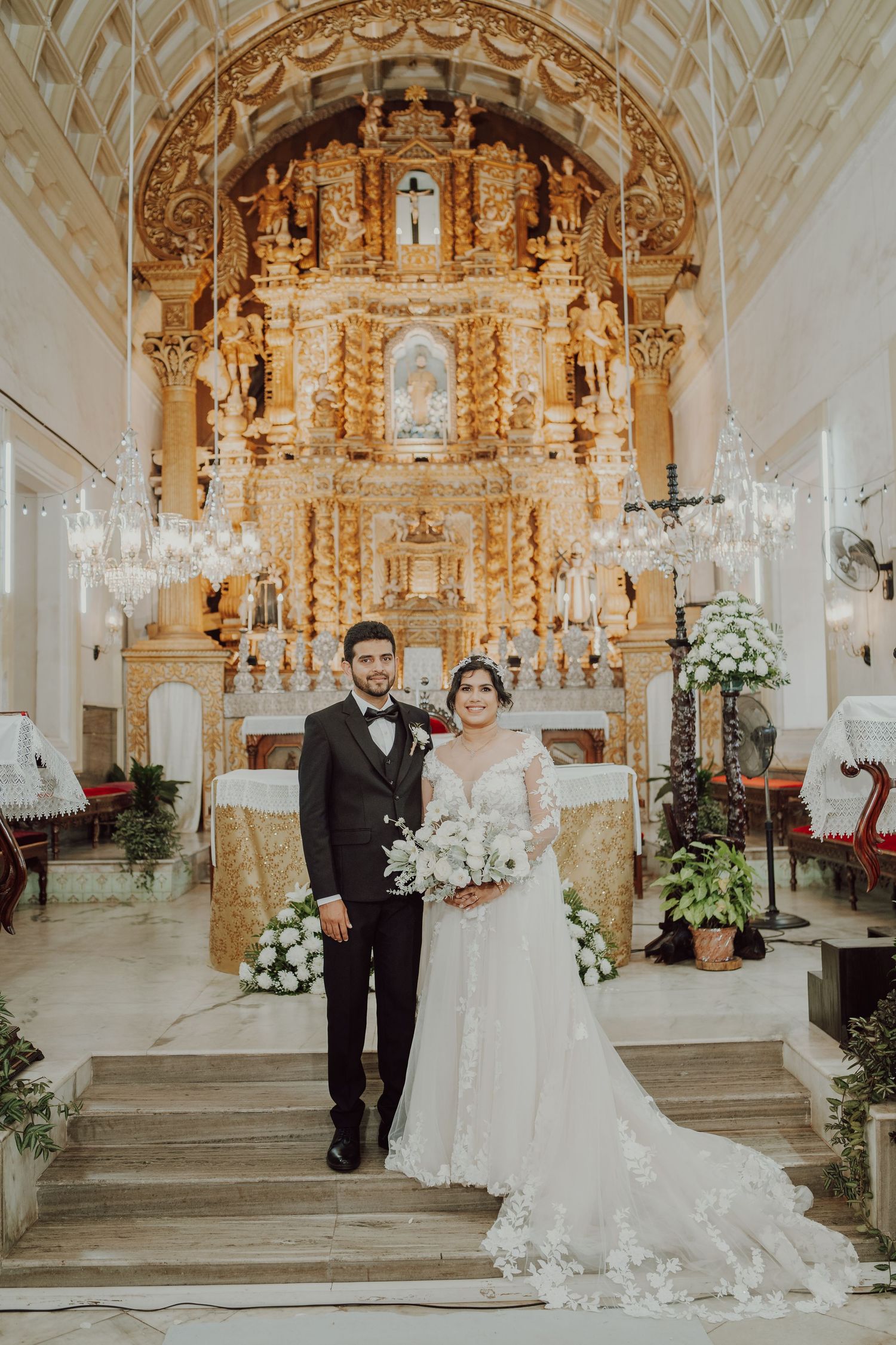 A wedding ceremony in an ornate Catholic church with baroque gold altarpiece and floral decorations.