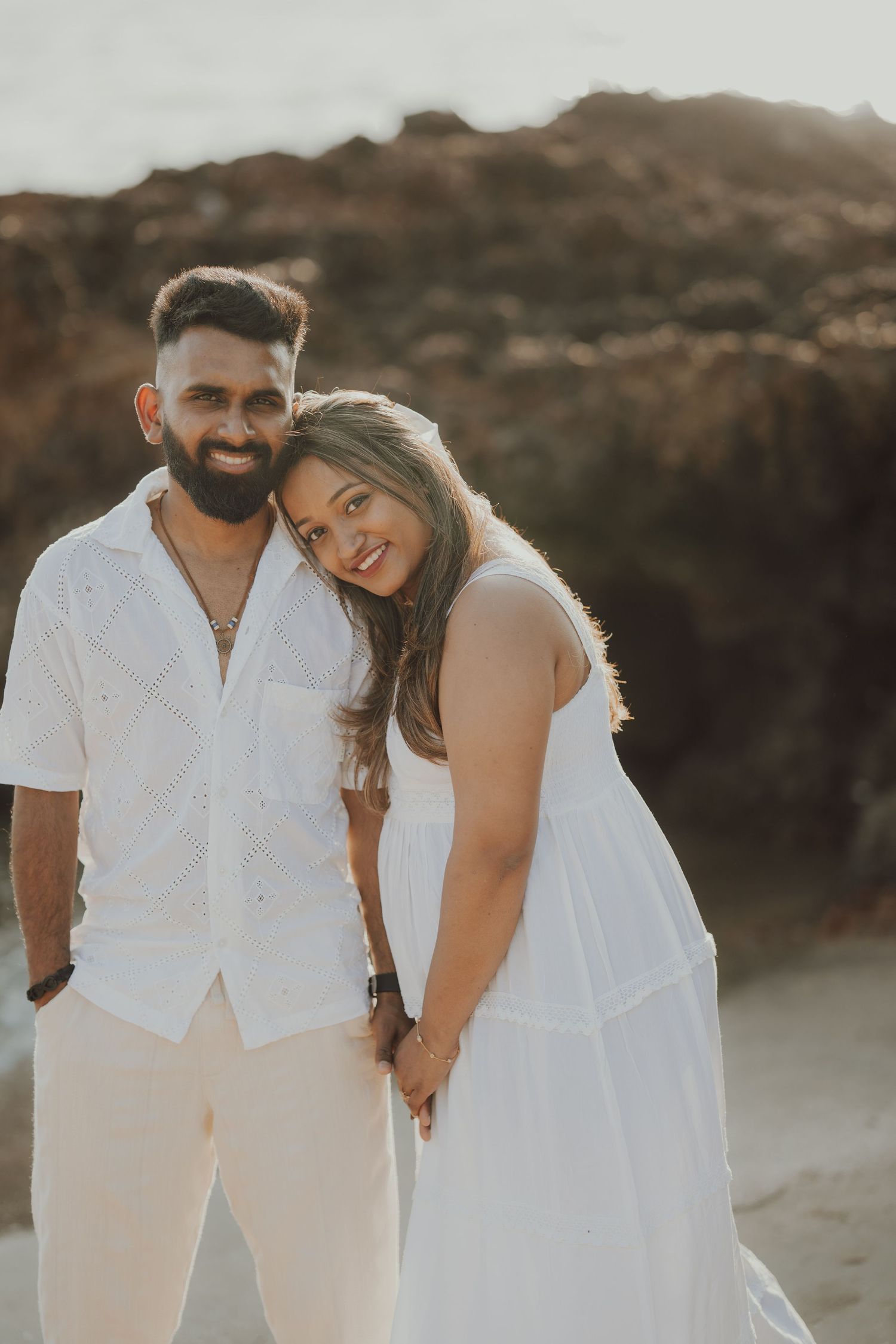 A couple in white outfits embraces lovingly on a beach during sunset with rocky cliffs in the background.