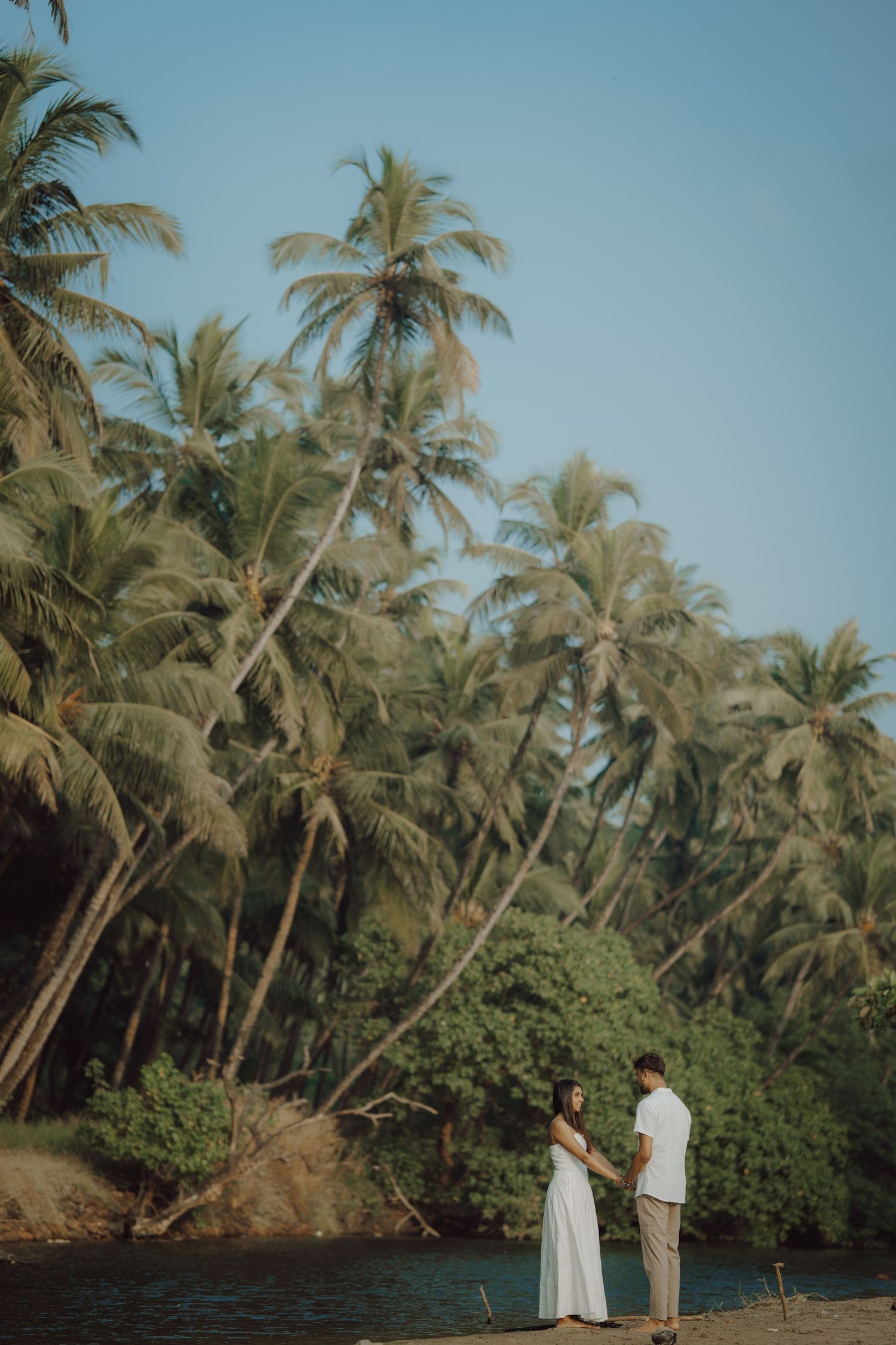 Couple in white clothing standing by a tropical lagoon surrounded by swaying palm trees on a sunny day.