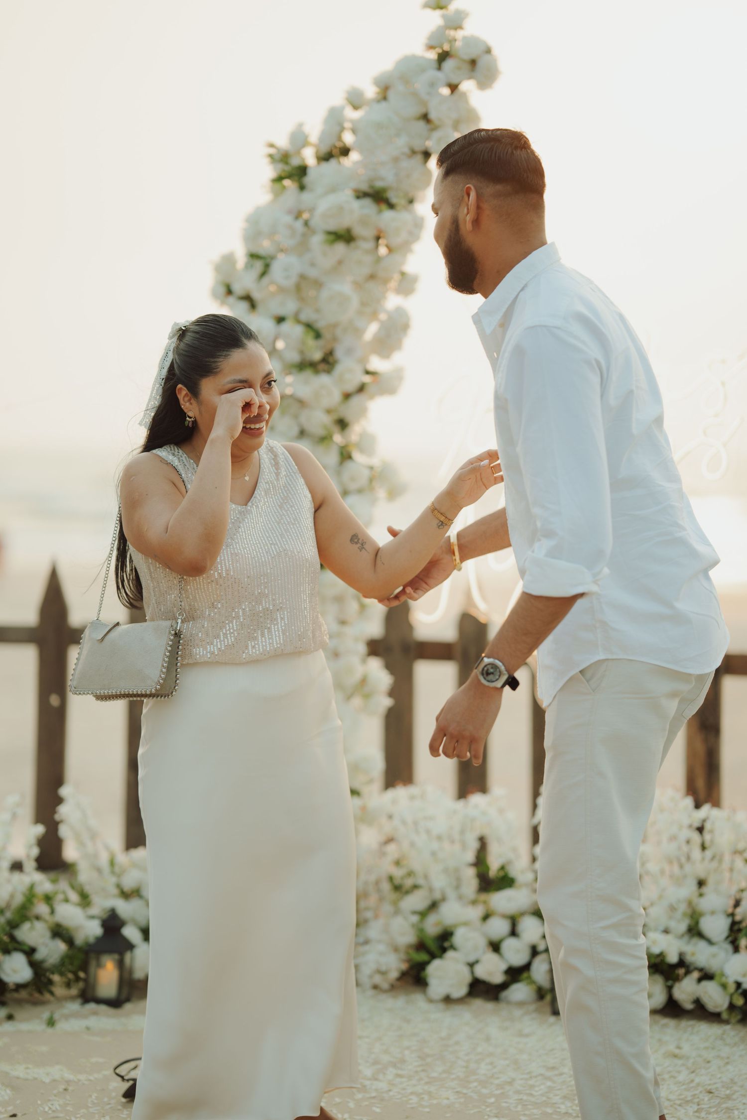 Emotional couple in white outfits during outdoor proposal near floral arch, woman wiping tears of joy.