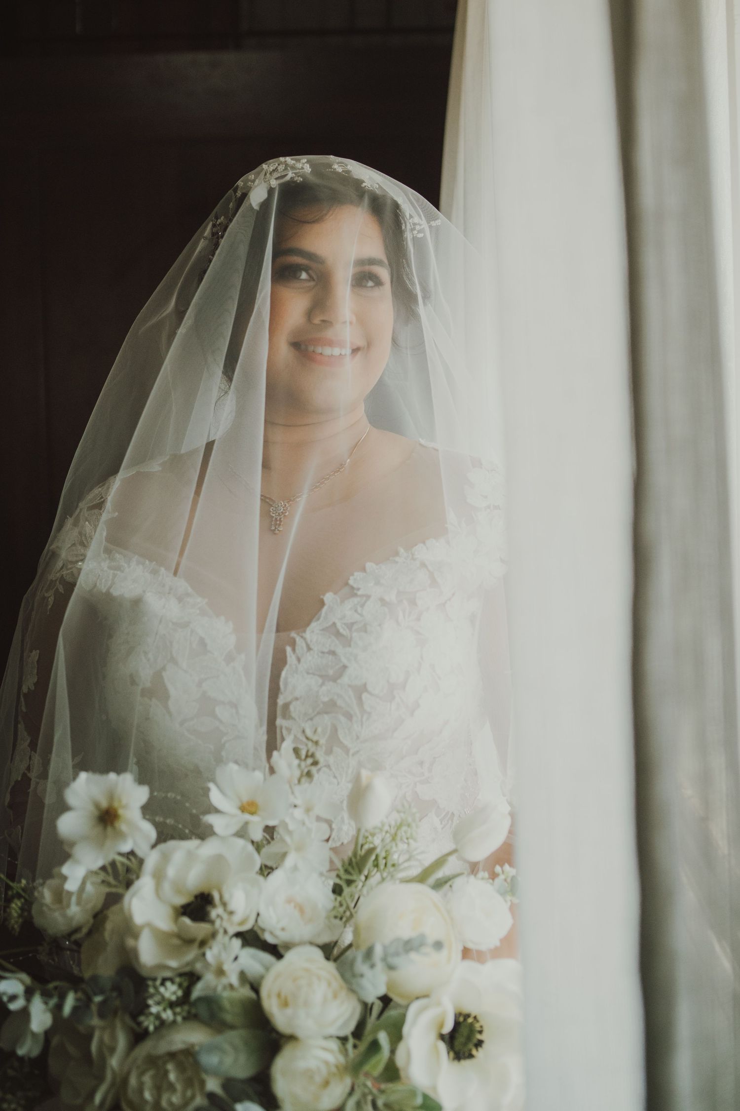 A radiant bridal portrait with white bouquet and flowing veil in natural window light.