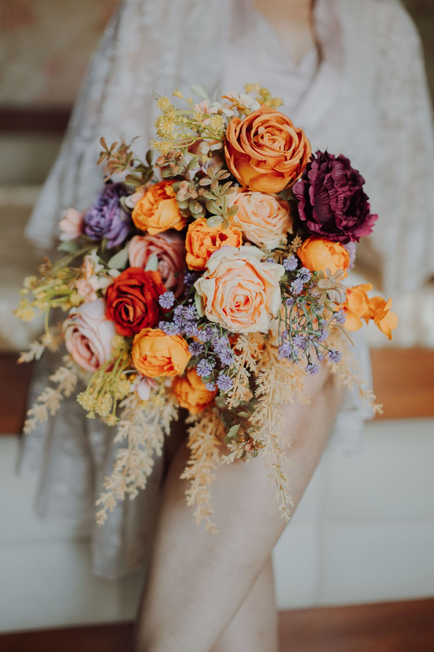 Bride in a lace gown holding a rustic wedding bouquet of orange roses, purple blooms and dried grasses.