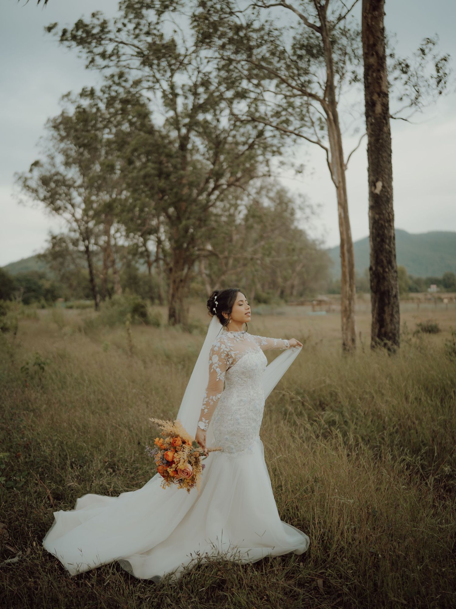 Bride in a lace mermaid wedding gown and veil holding a bouquet in a grassy field with trees and hills.