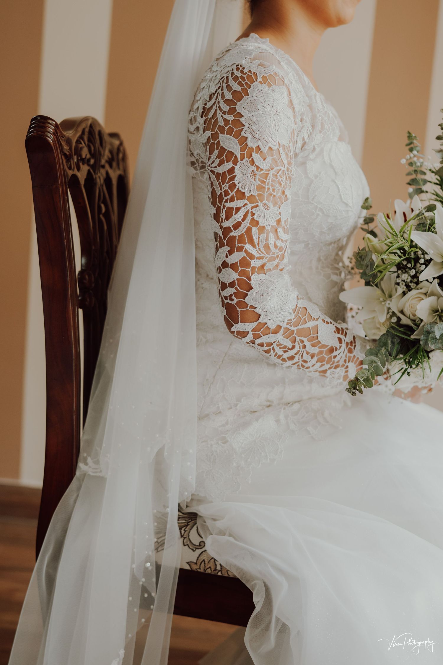 A detailed lace sleeve wedding dress with a flowing veil is elegantly draped over an antique wooden chair with floral bouquet.