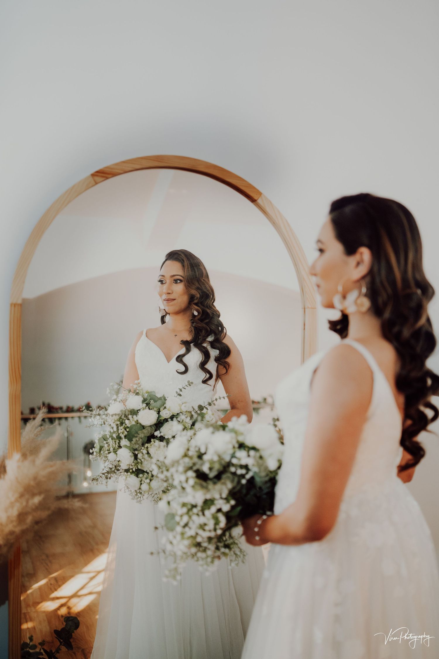 Bride holding white floral bouquet gazes at her reflection in an arched mirror during wedding preparations.