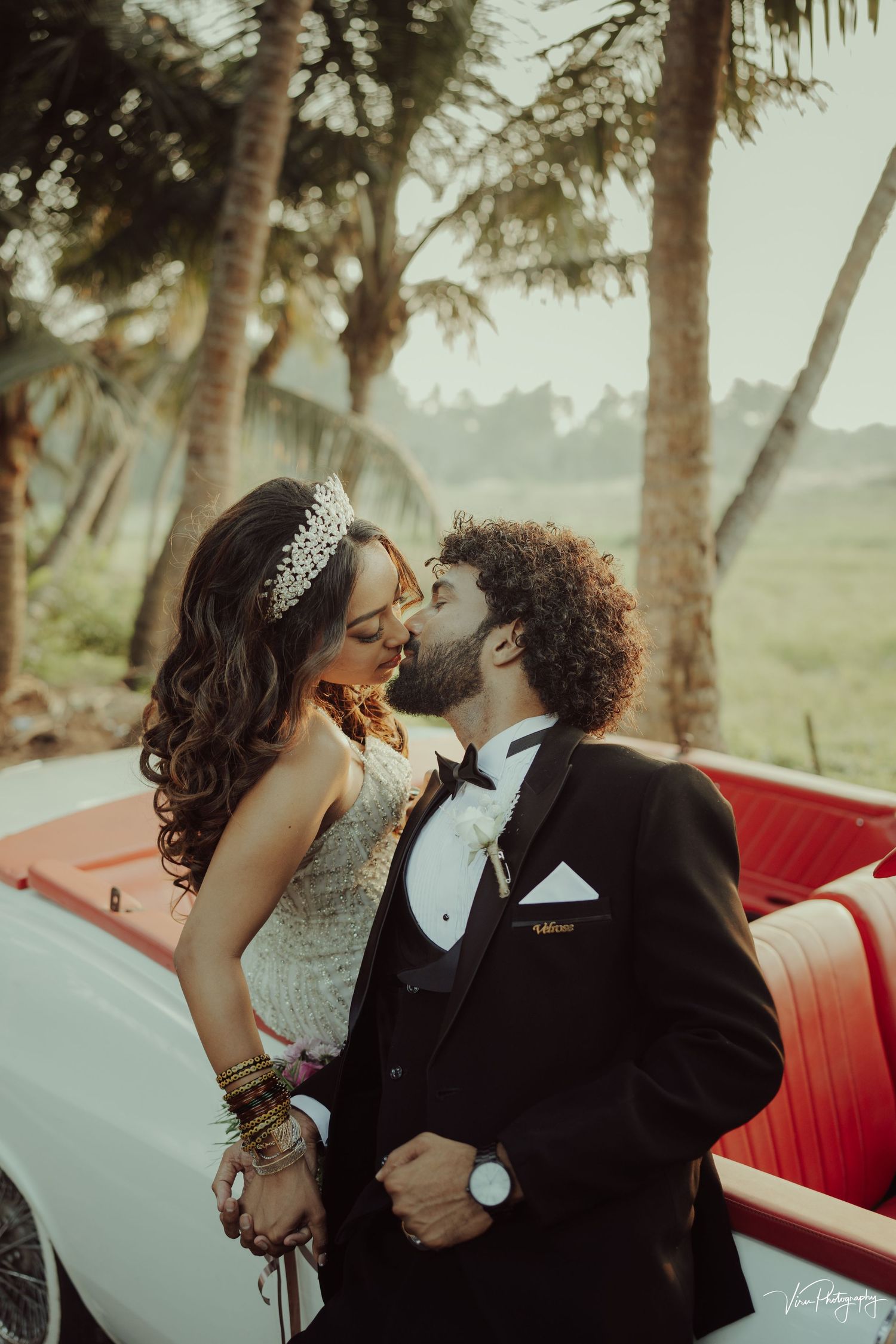 Romantic couple in formal wear share an intimate moment under palm trees near a vintage car.
