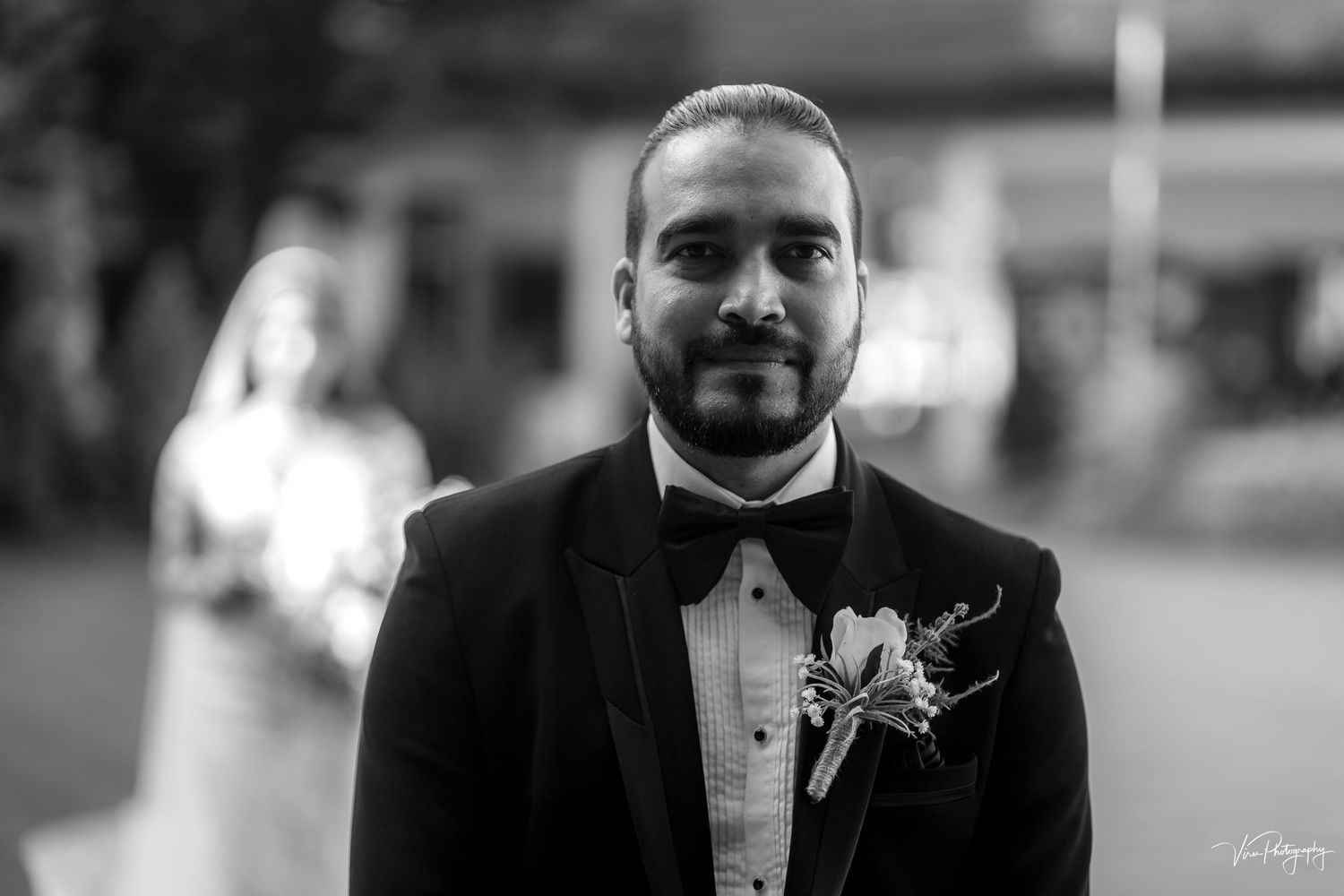 Black and white portrait of groom in tuxedo with boutonniere at outdoor wedding ceremony.