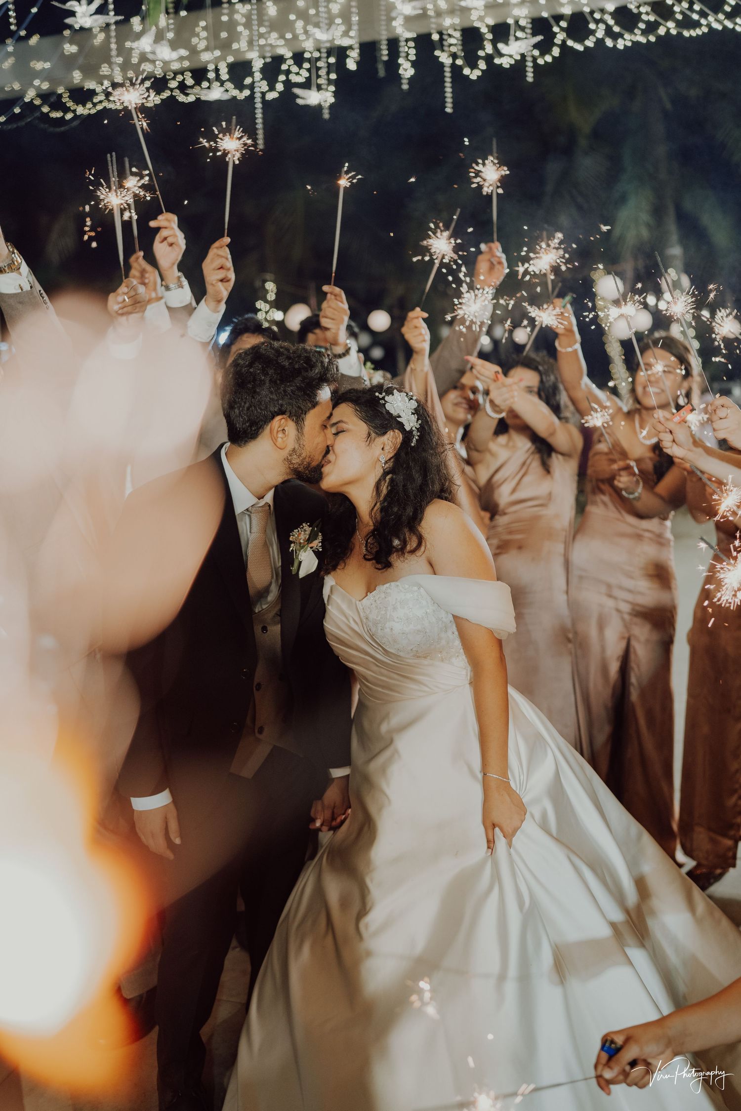 Wedding couple shares romantic kiss under sparklers held by wedding party guests at nighttime reception celebration.