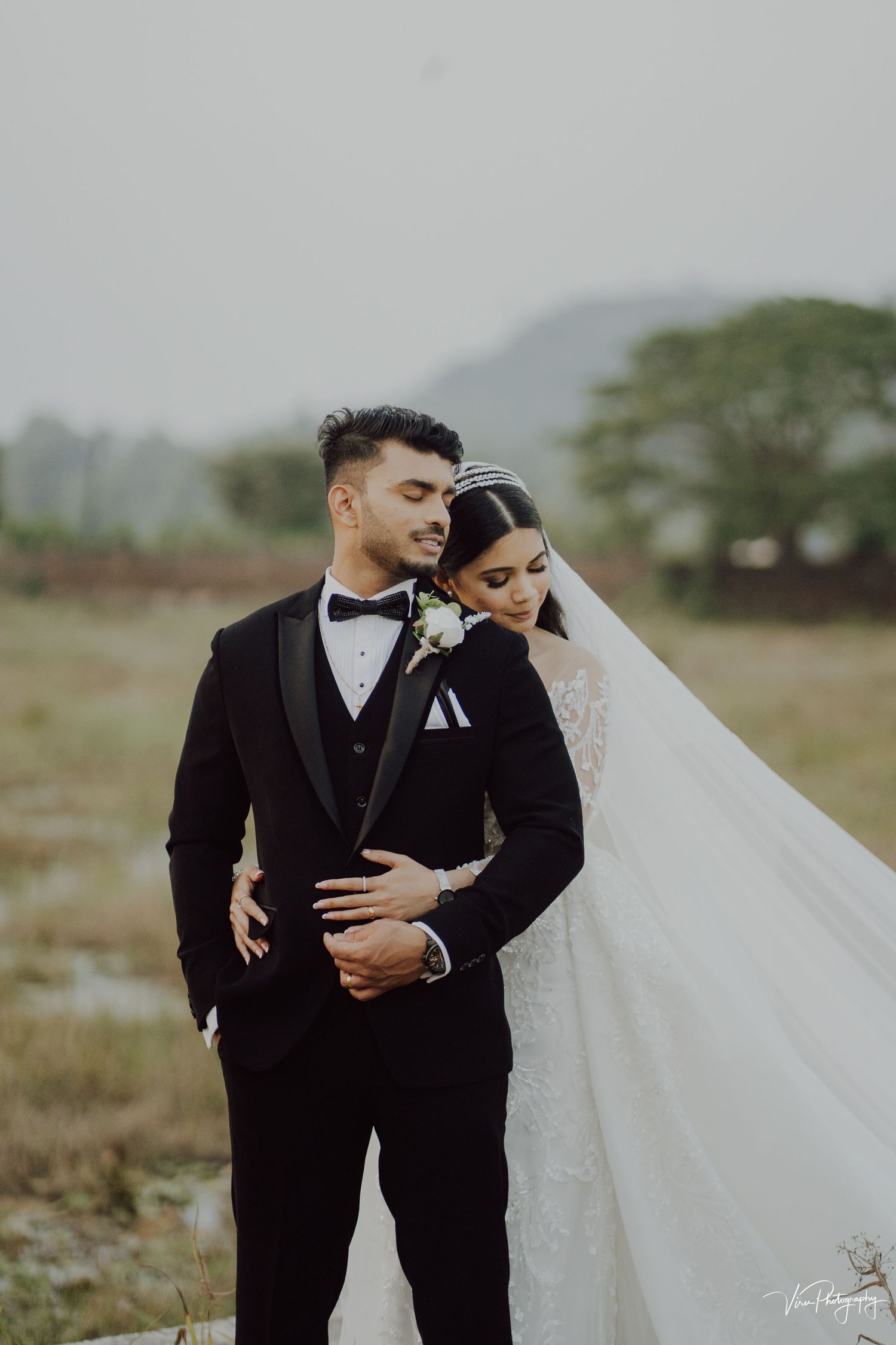 A bride and groom embrace outdoors in formal wedding attire against a misty mountain backdrop.
