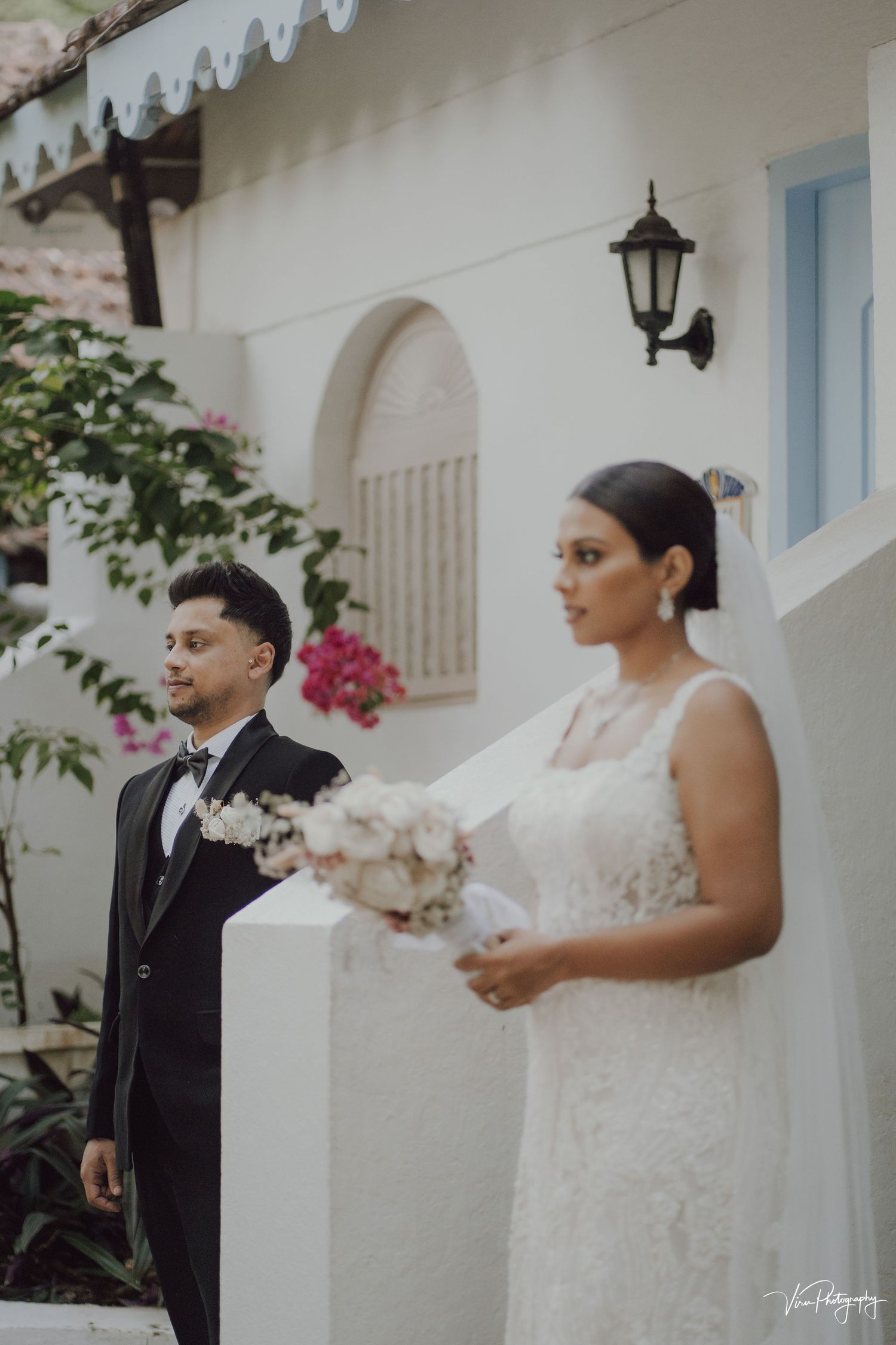 Wedding couple in elegant attire stand outside Mediterranean-style building with white walls and decorative lantern.