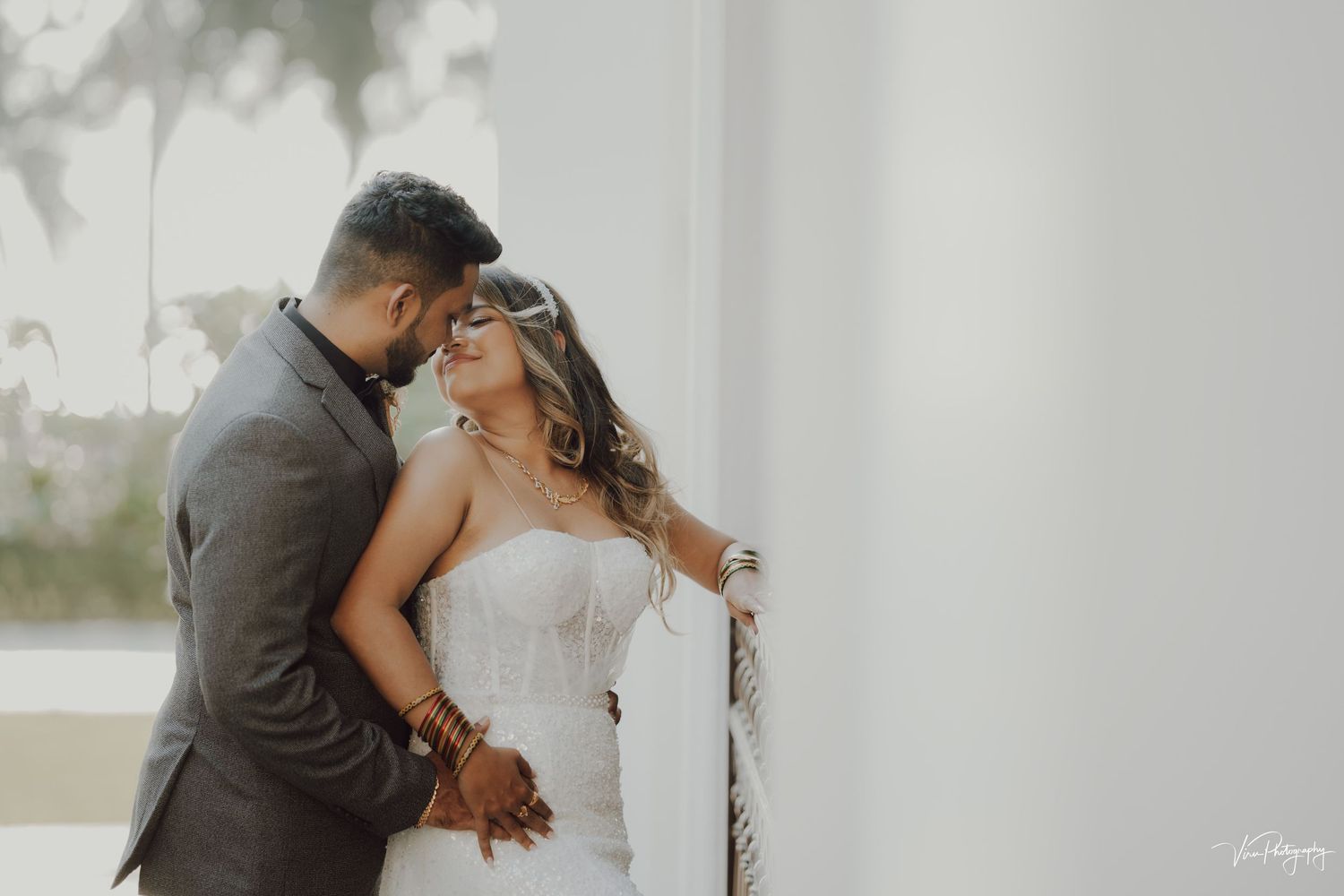 A romantic couple in wedding attire embrace intimately against a white wall in soft natural light.