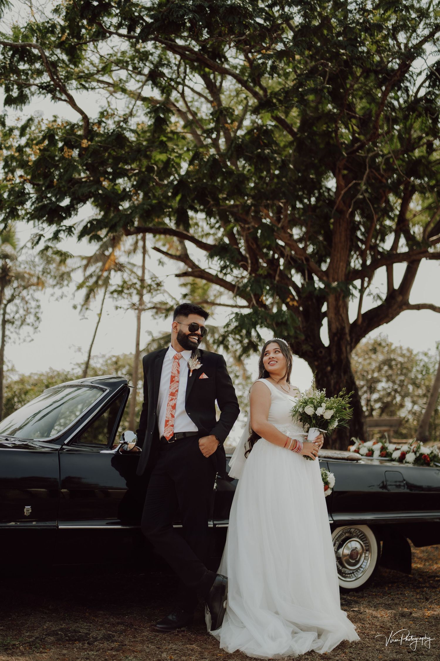 Wedding couple poses by vintage black car under large tree in romantic outdoor setting.