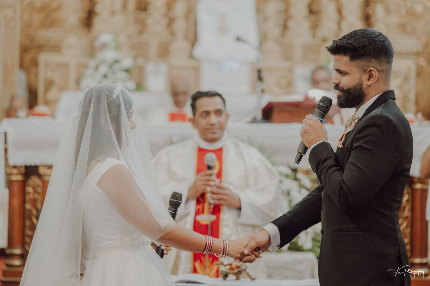 During a traditional Catholic church ceremony, a couple holds hands at the altar while a priest officiates their wedding.