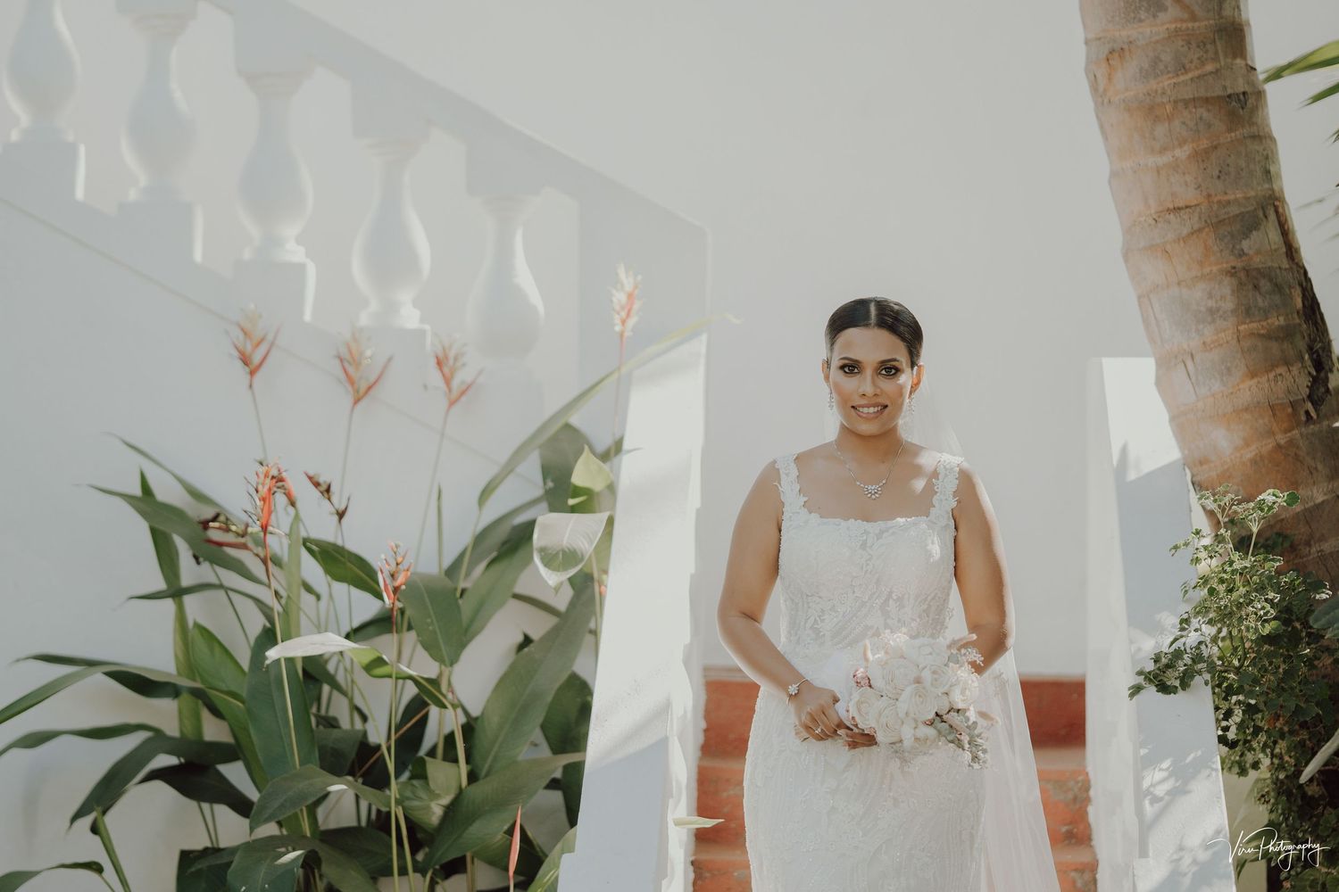 A bride in a classic white wedding dress stands elegantly between palm trees with a tropical backdrop.