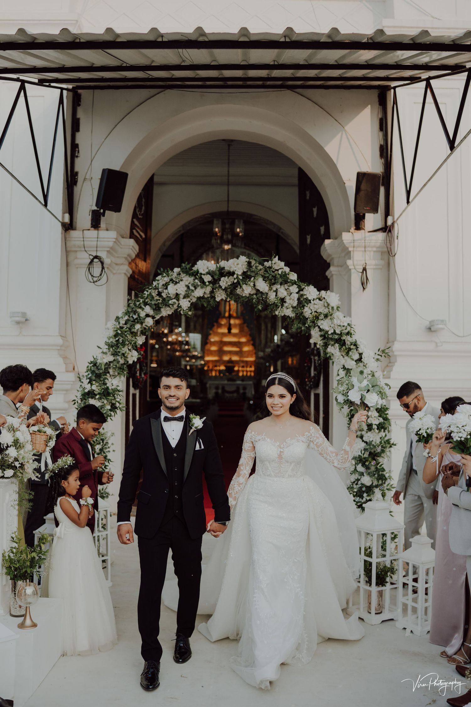 Newlyweds exit church under white floral arch while guests line the walkway at elegant wedding ceremony.