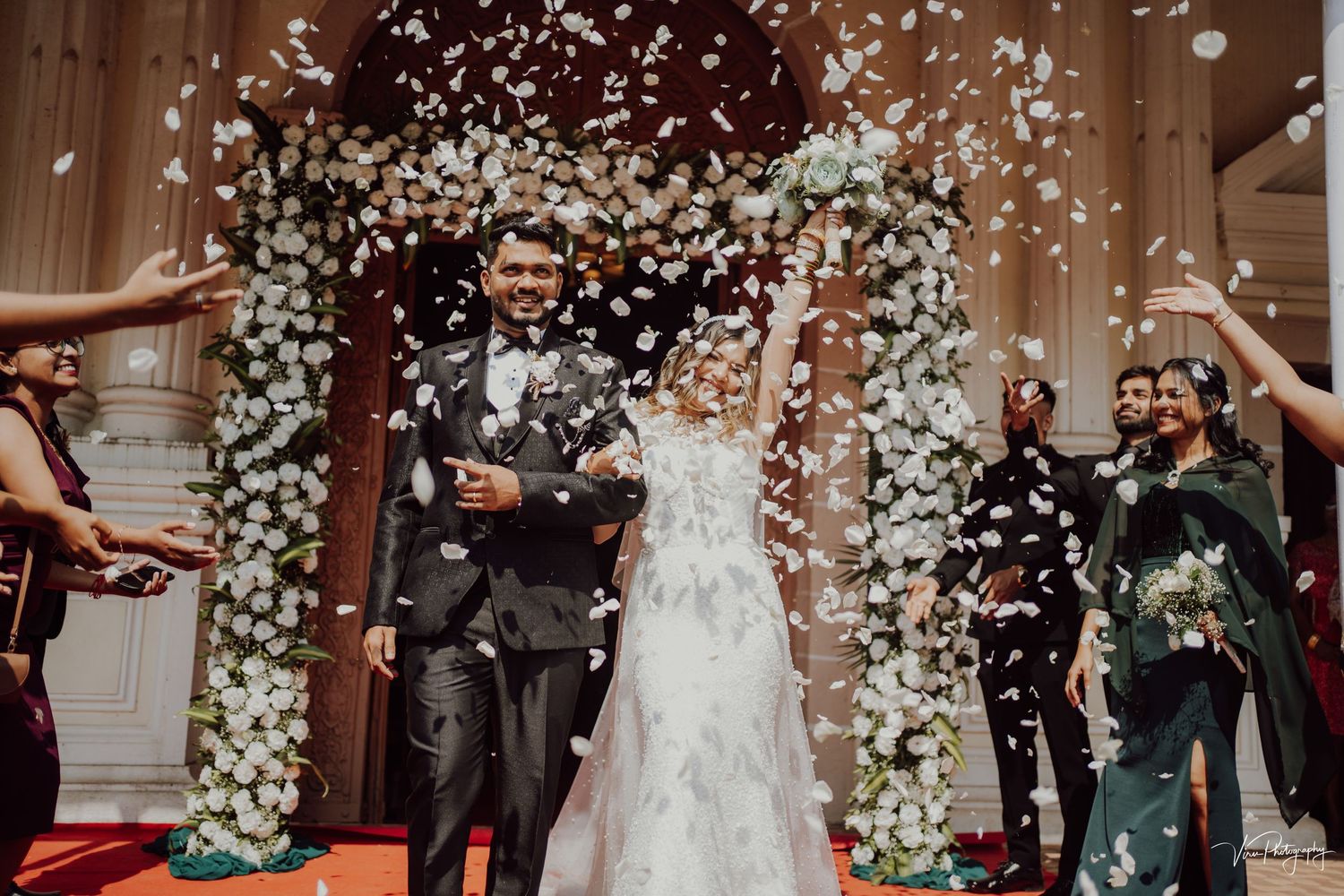 Newlyweds walk under floral arch while guests shower them with white petals during joyful wedding ceremony exit.