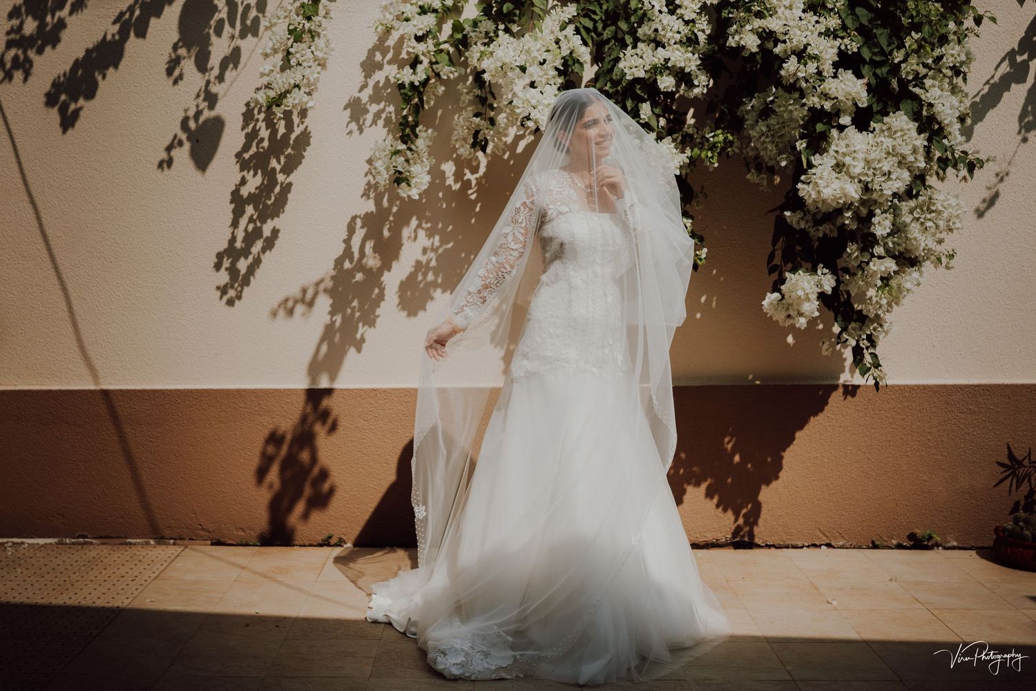 A flowing white wedding gown and veil cast elegant shadows against a beige wall beneath overhanging foliage.