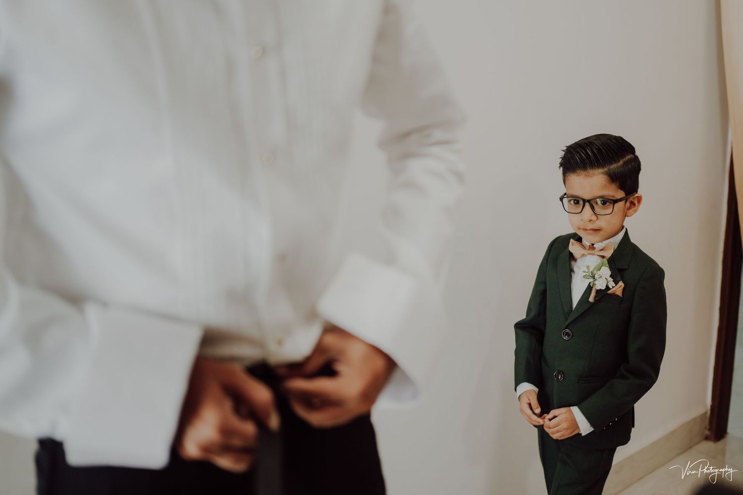 A small figure wearing a dark suit and glasses stands against a white wall while someone adjusts clothing in the foreground.
