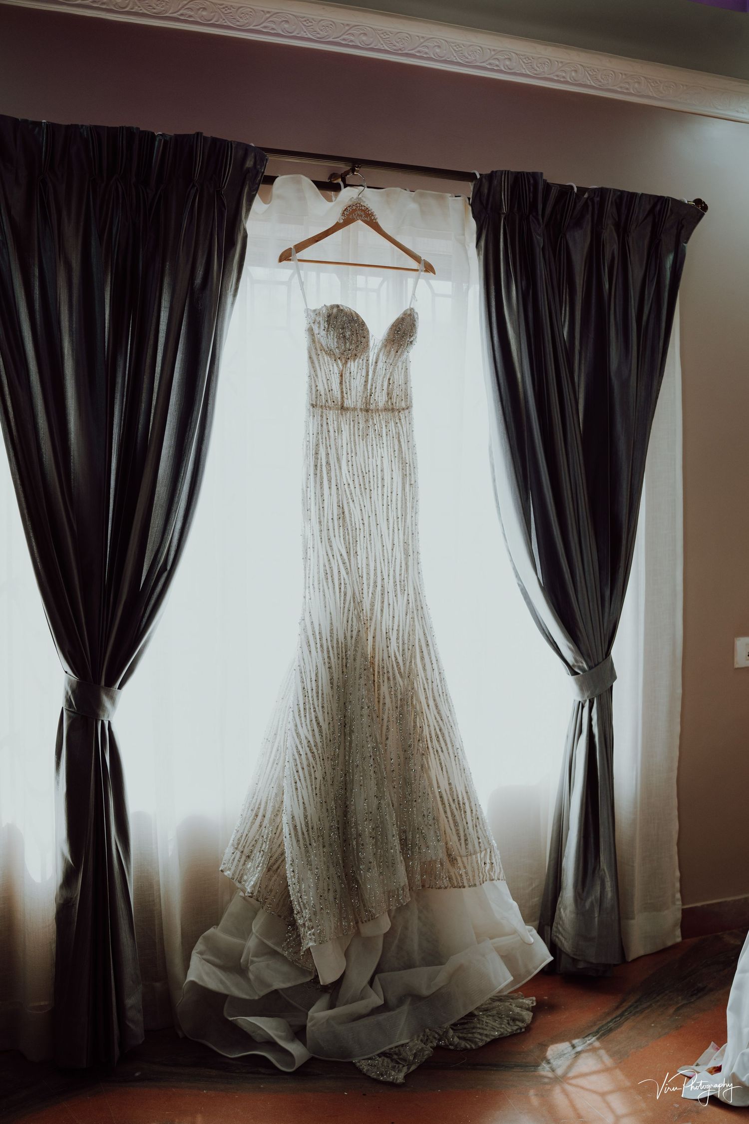An elegant white lace wedding gown hangs from a wooden hanger between dark curtains against a bright window.