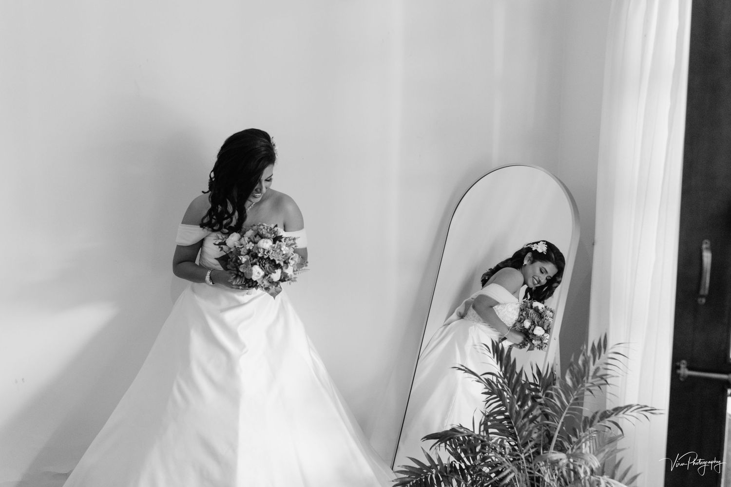 A black and white wedding photograph showing a bride with a bouquet looking at her reflection in a full-length mirror.