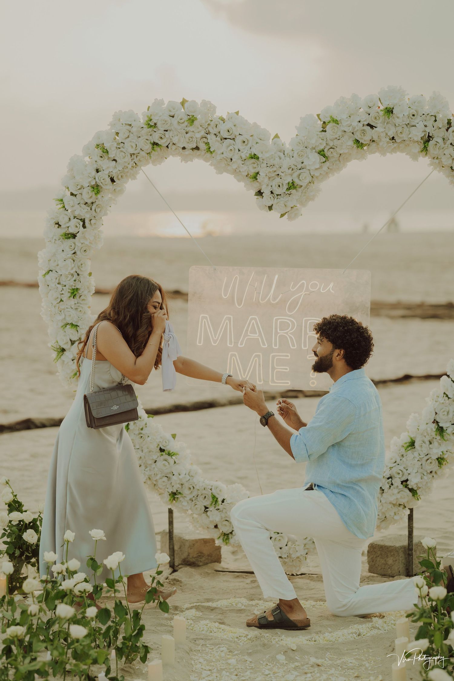 A romantic beach proposal during sunset under a beautiful white floral arch.