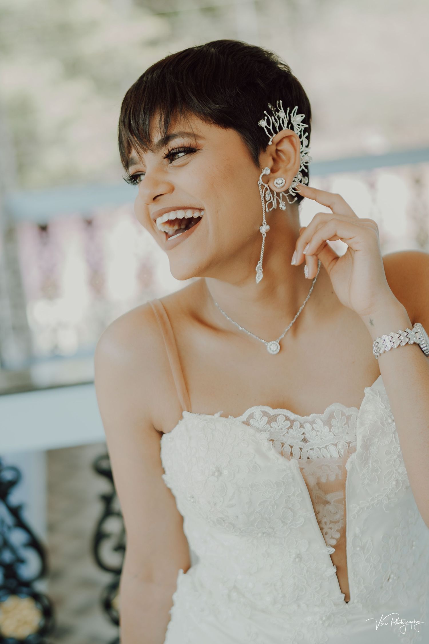 A bride wearing a strapless lace wedding dress smiles joyfully while adjusting her crystal ear cuff and jewelry.