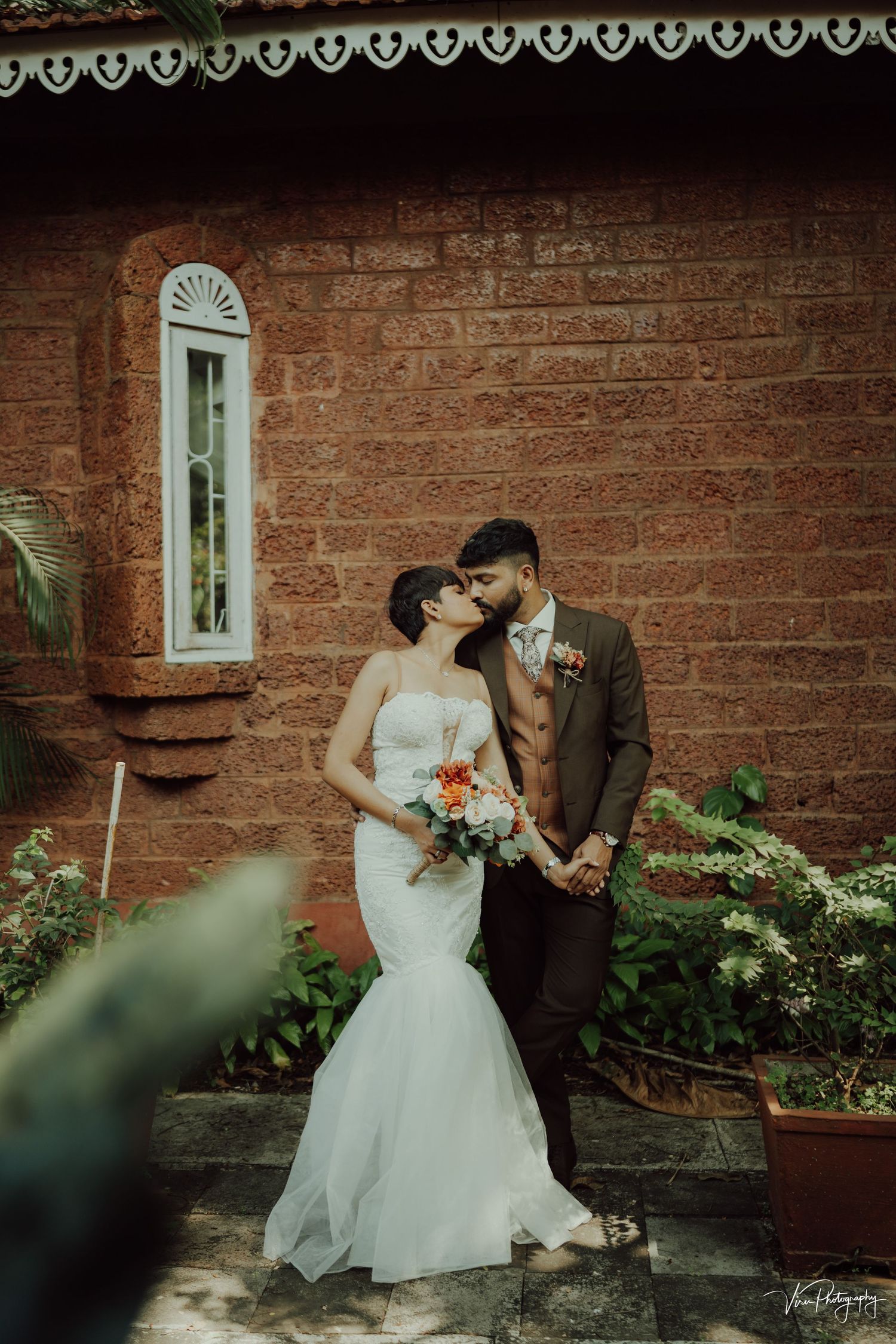 A romantic wedding couple poses against a rustic brick wall with an ornate window and potted plants framing the scene.