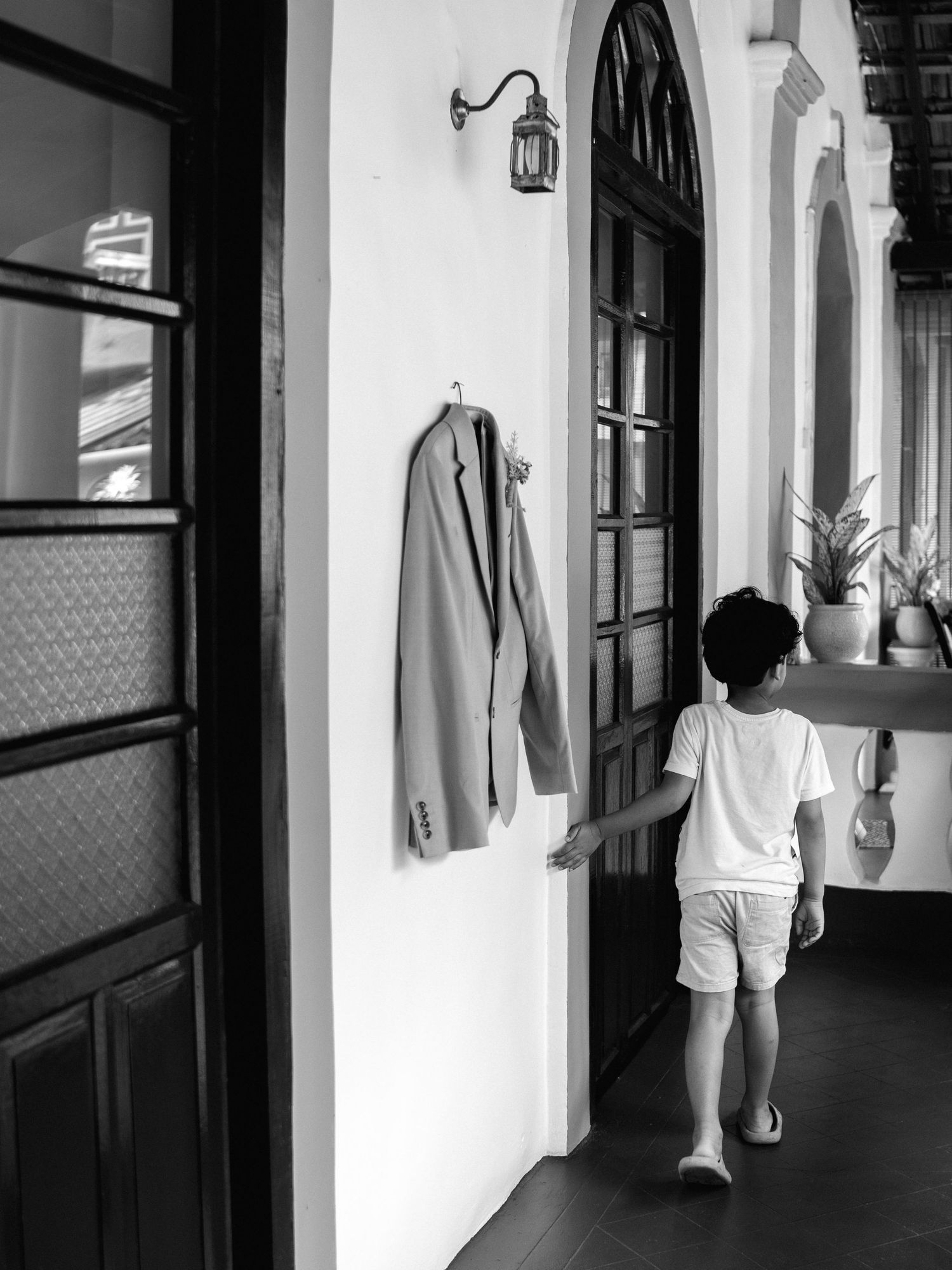 Black and white photo of a young boy walking down a sunlit arched corridor past a blazer hanging on the wall beside a vintage lantern.