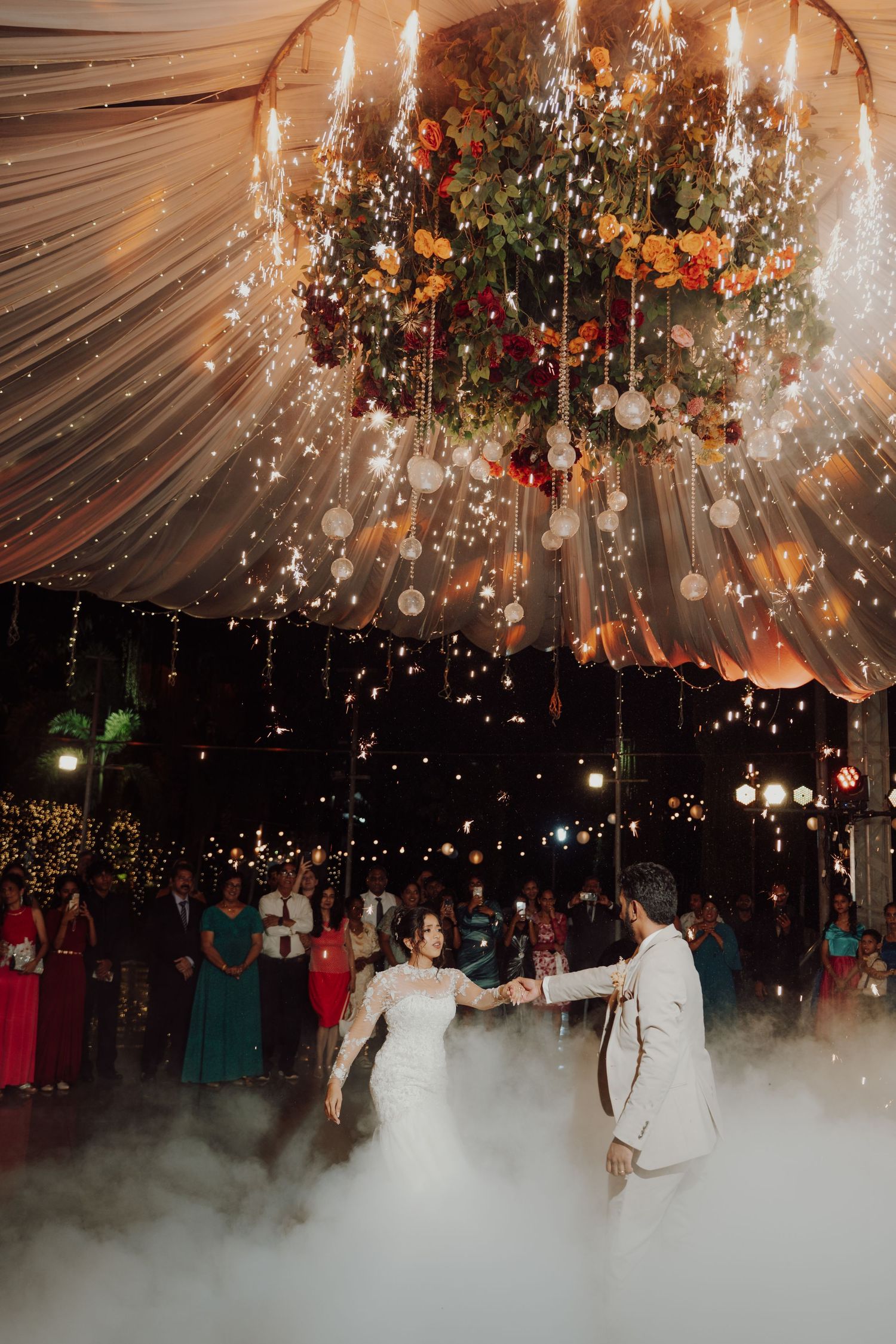 A wedding couple shares a first dance in misty fog beneath a sparkling floral chandelier and draped fabric ceiling at night.