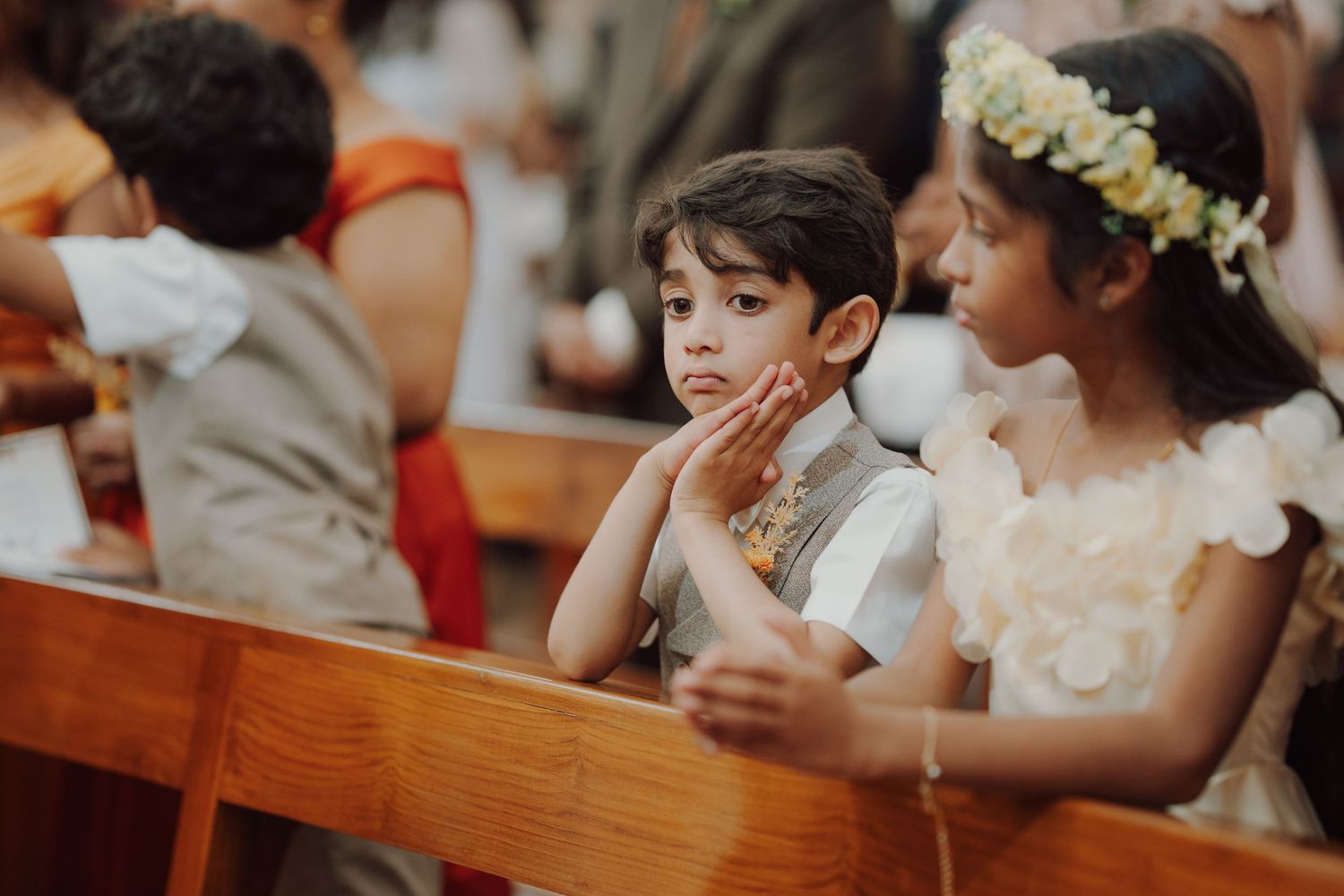 A young boy rests his chin on his hand beside a flower girl with a floral crown as they sit on a wooden church pew.