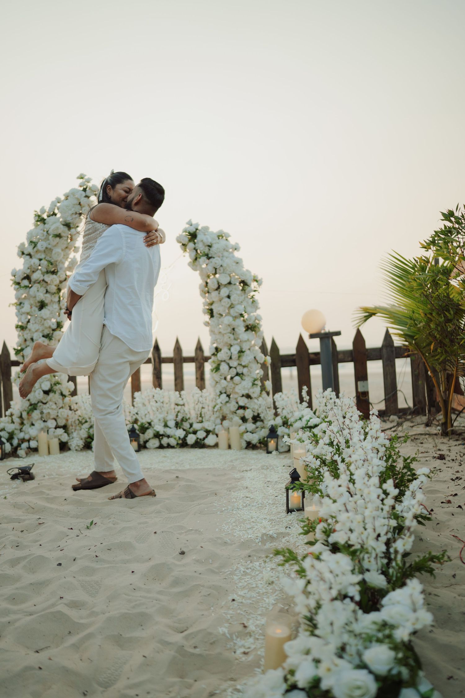 Couple embracing under white floral arch on sandy beach at sunset with candles and flowers.
