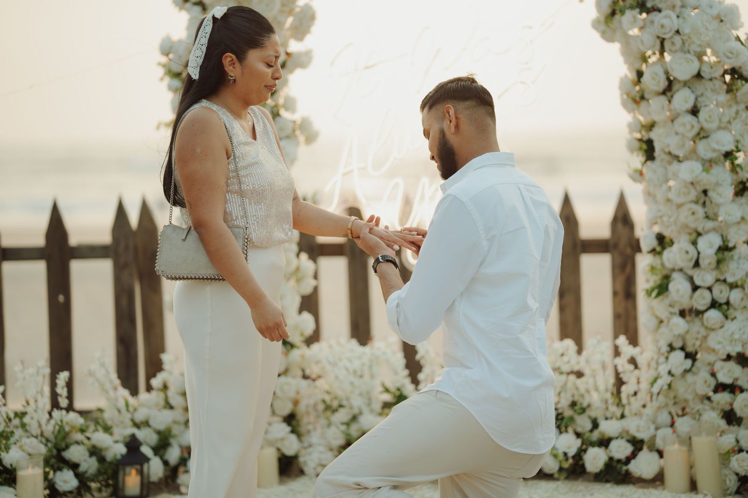Man proposes to woman on beach at sunset surrounded by white floral arrangements and string lights.