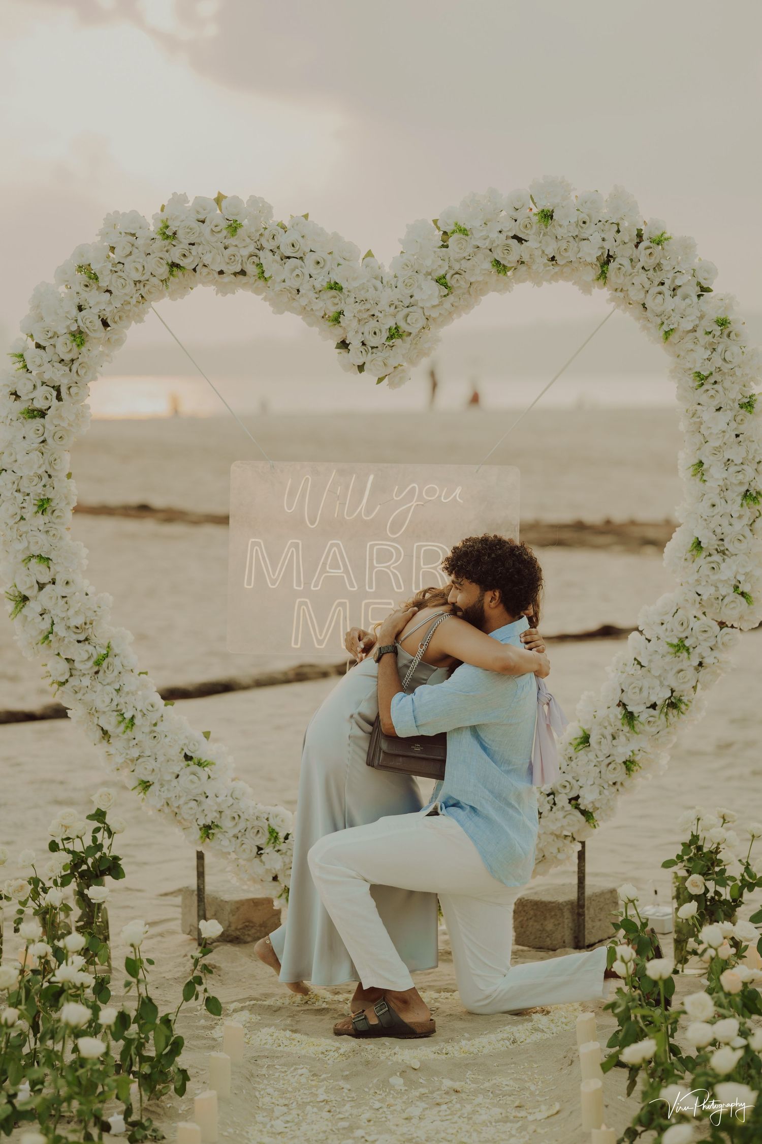 A romantic beach proposal scene under a heart-shaped floral arch with white flowers at sunset.