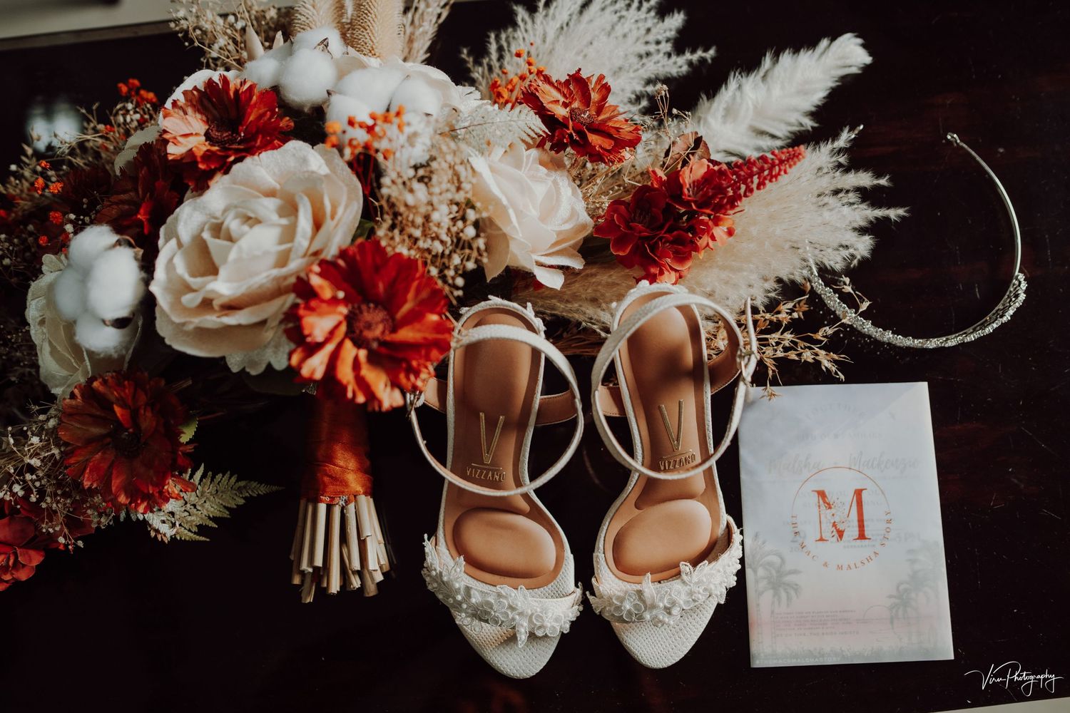Wedding details featuring white bridal shoes, red and white floral bouquet, and invitation card with M monogram against dark background.