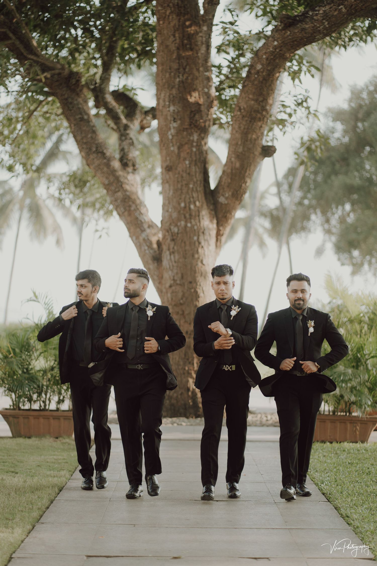 A group in matching black suits and boutonnieres walk together outside under a large tree during a wedding event.