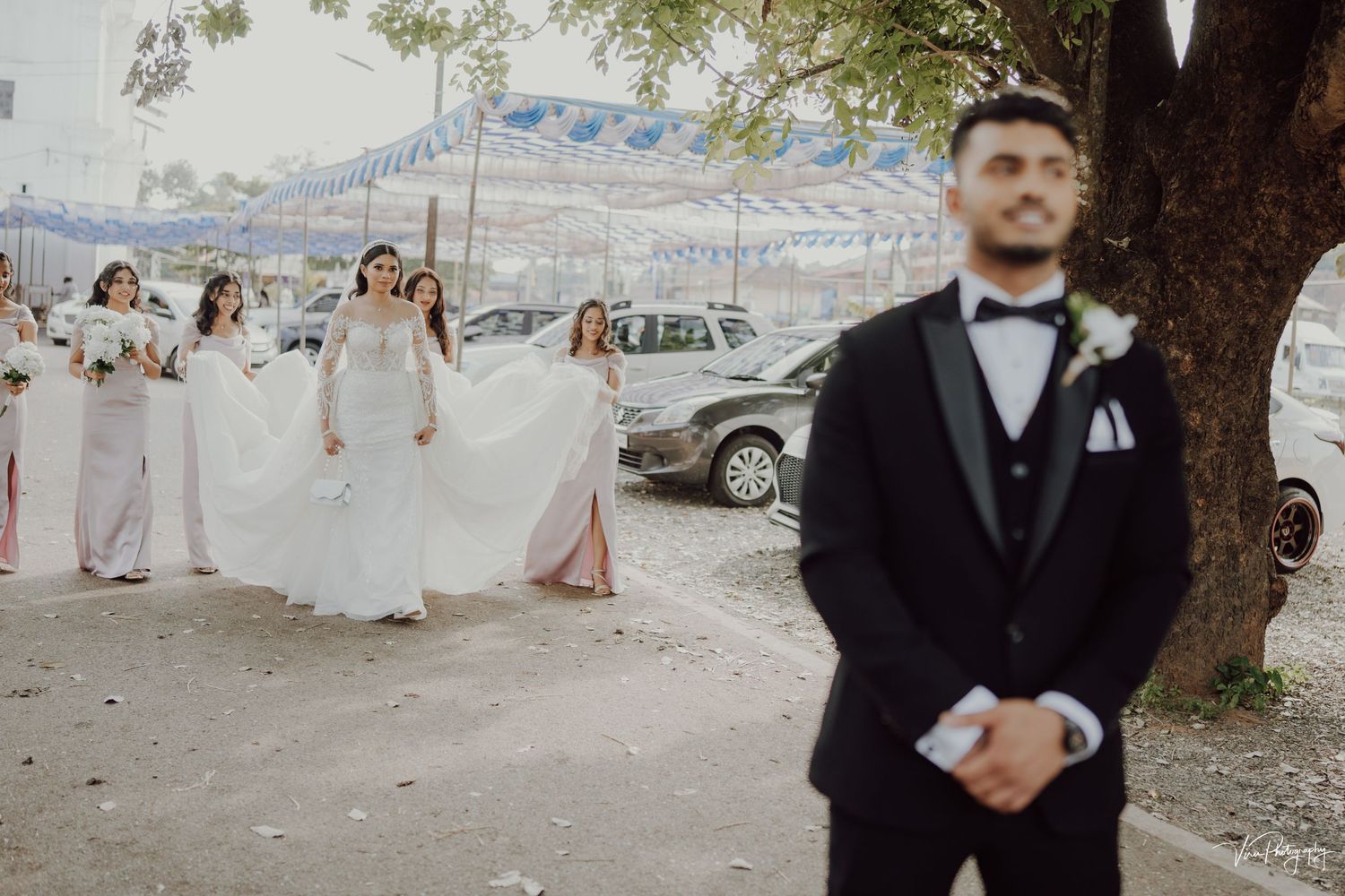 Wedding party gathers outside with bridesmaids in pink dresses walking behind a groom in a black tuxedo near a tree.