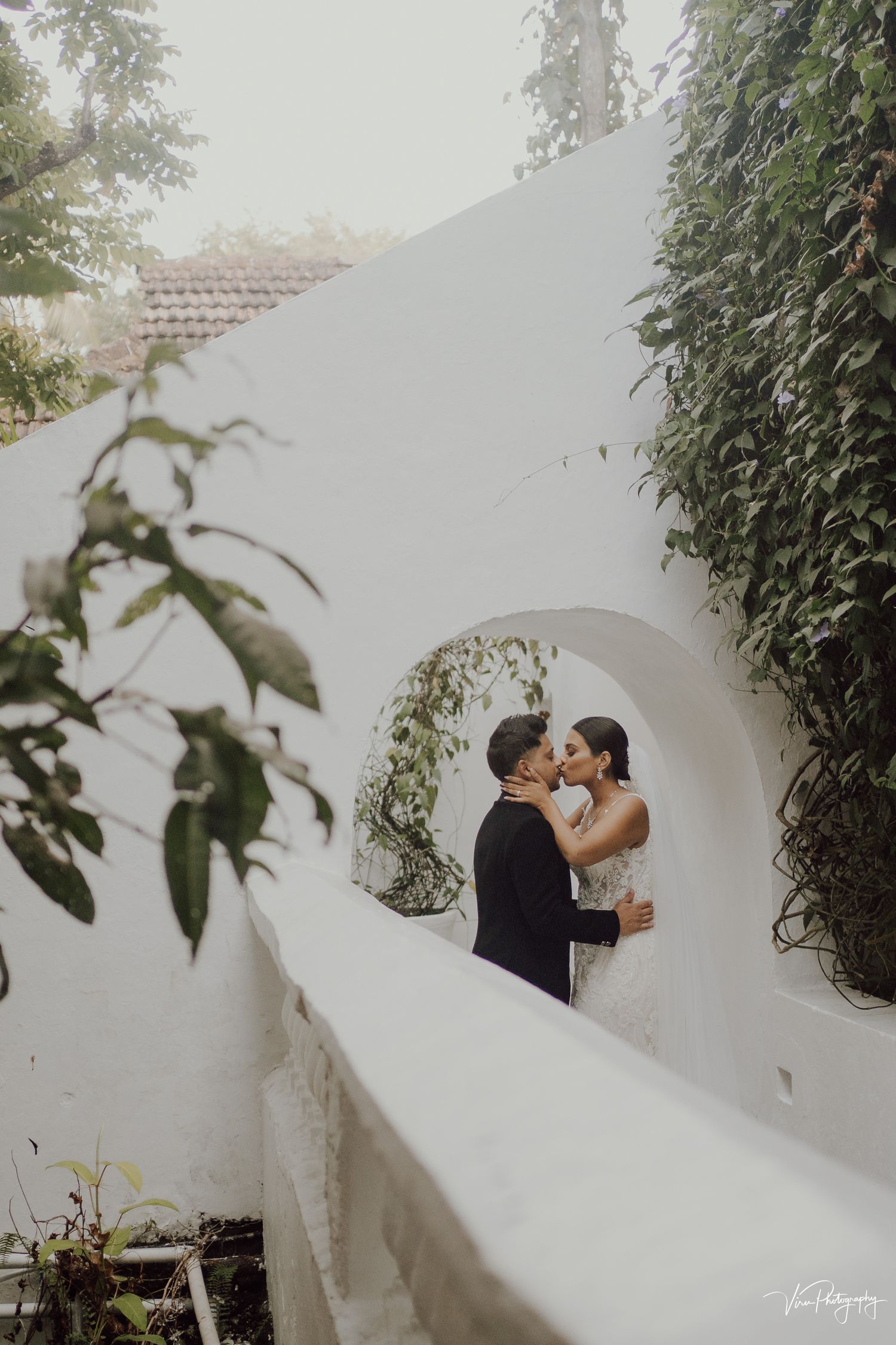 A romantic couple embraces in a white Mediterranean-style archway framed by lush greenery.