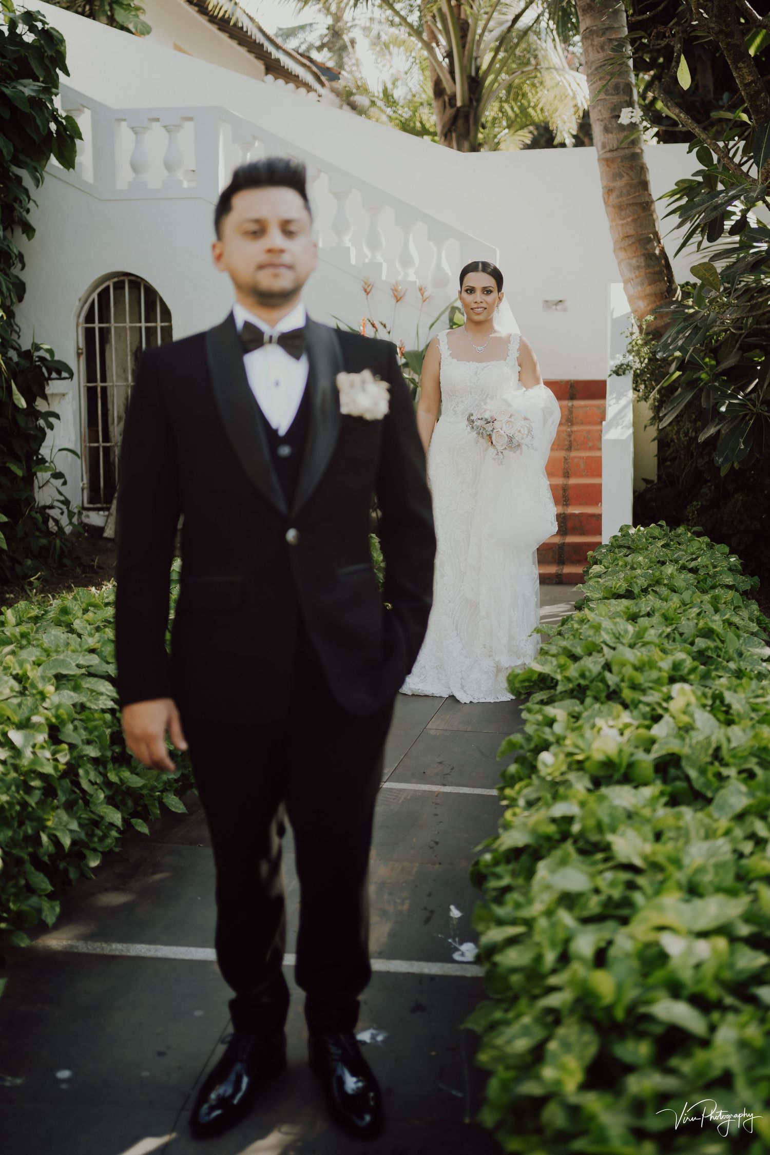 A traditional wedding scene in a garden courtyard with the bride in white and groom in black tuxedo standing on stone steps.