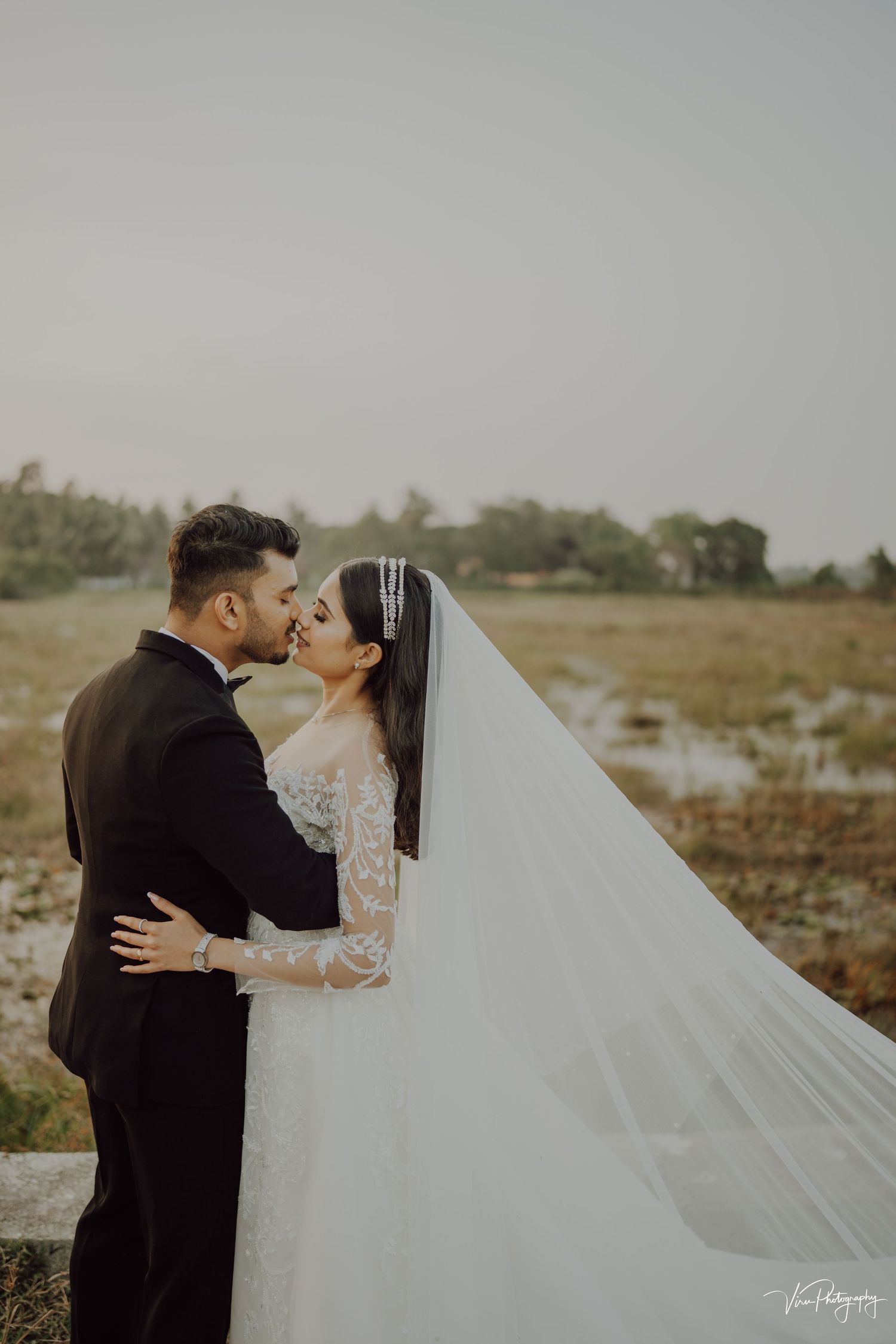 A couple shares a romantic kiss in a field at sunset, with the bride wearing a long veil and lace sleeve wedding gown.