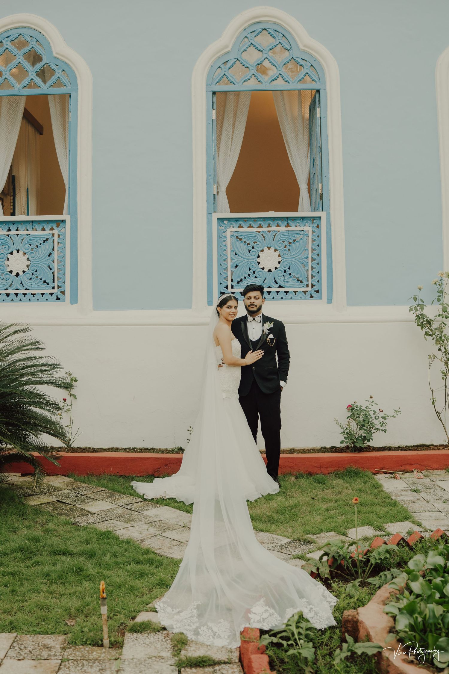 Couple poses outside ornate building with blue Moroccan-style windows and flowing white wedding dress train on green lawn.