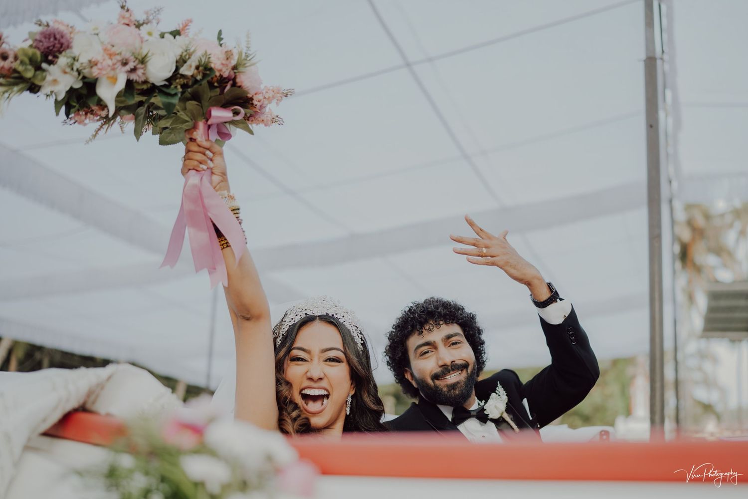 A joyful couple celebrates at an outdoor wedding ceremony while raising bouquets and arms under a white tent.