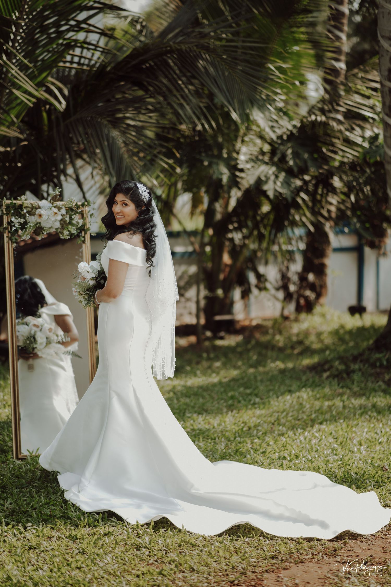 A bride in an elegant off-shoulder white mermaid wedding gown with long train poses outdoors among tropical palm trees.