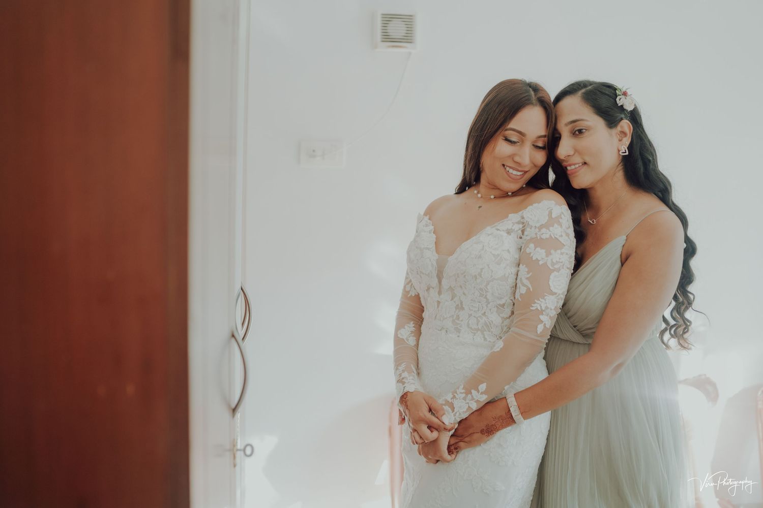 Two women in elegant formal dresses share a tender moment together against a simple white wall interior.