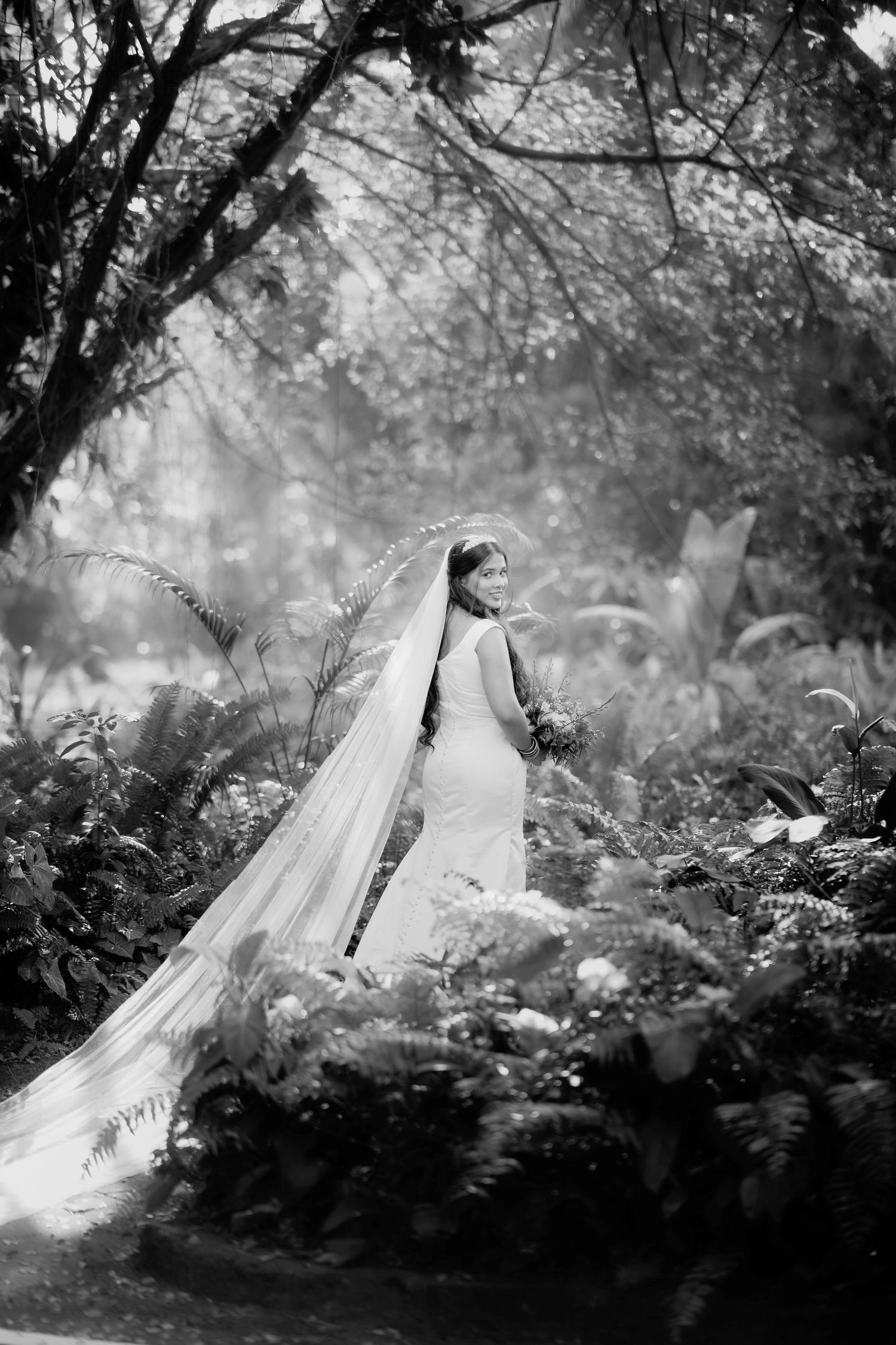 A dreamy black and white bridal portrait showing a flowing long veil amid a misty forest garden setting.
