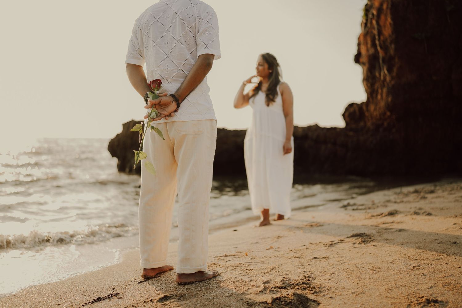 Couple walking together on sandy beach at sunset with cliffs and ocean waves in background.
