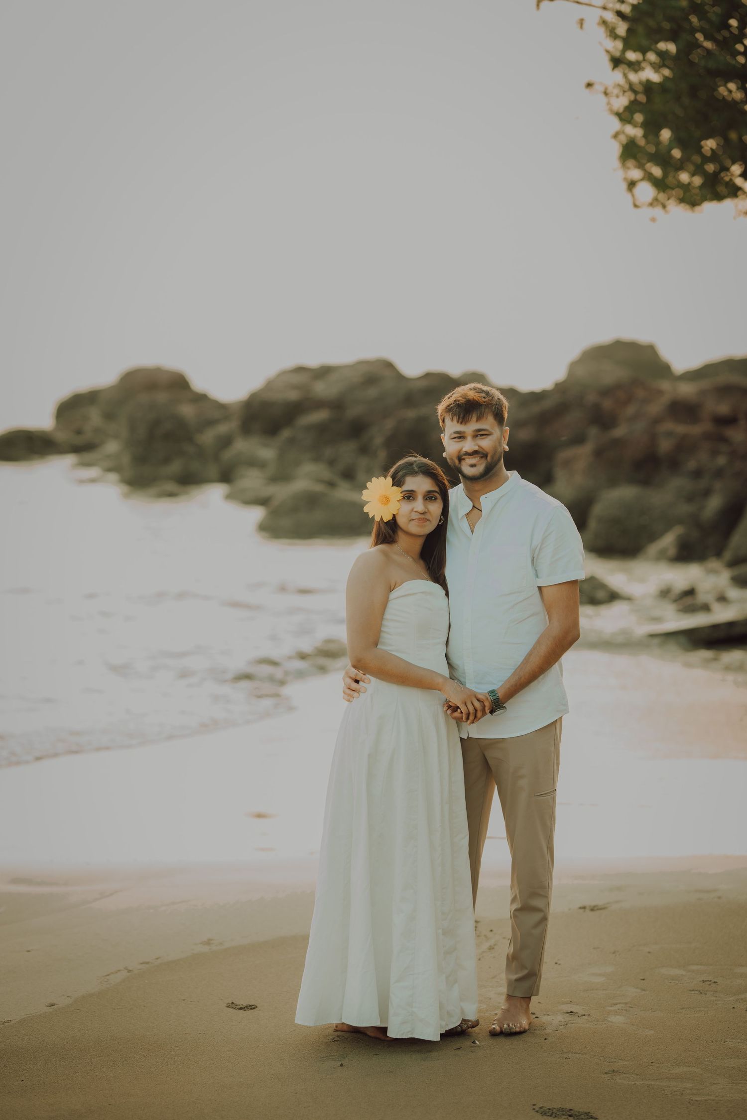A couple embraces on a sandy beach at sunset with rocky outcrops visible in the background.