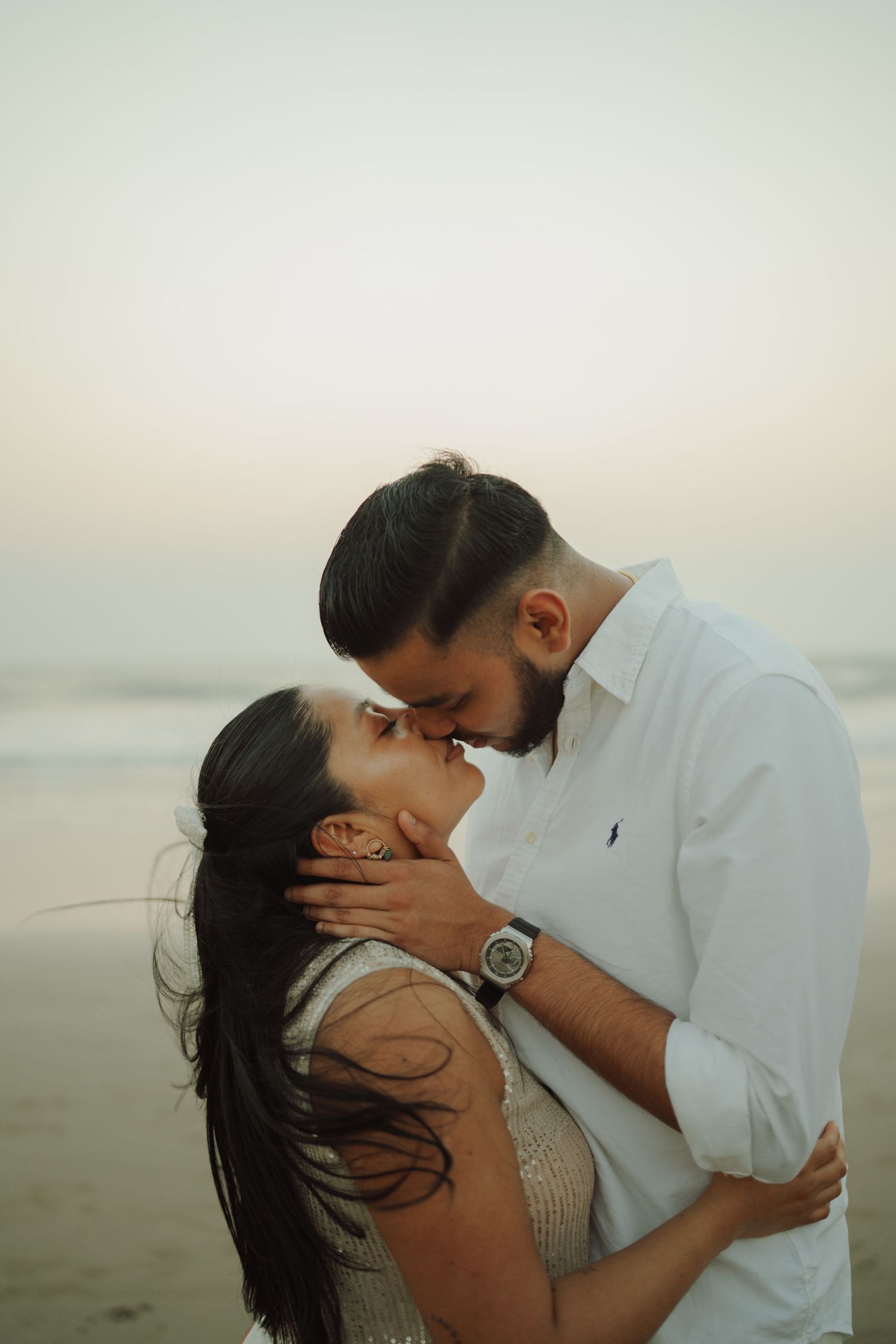 Couple sharing a romantic kiss on a beach at sunset, man in white shirt holding woman's face tenderly.